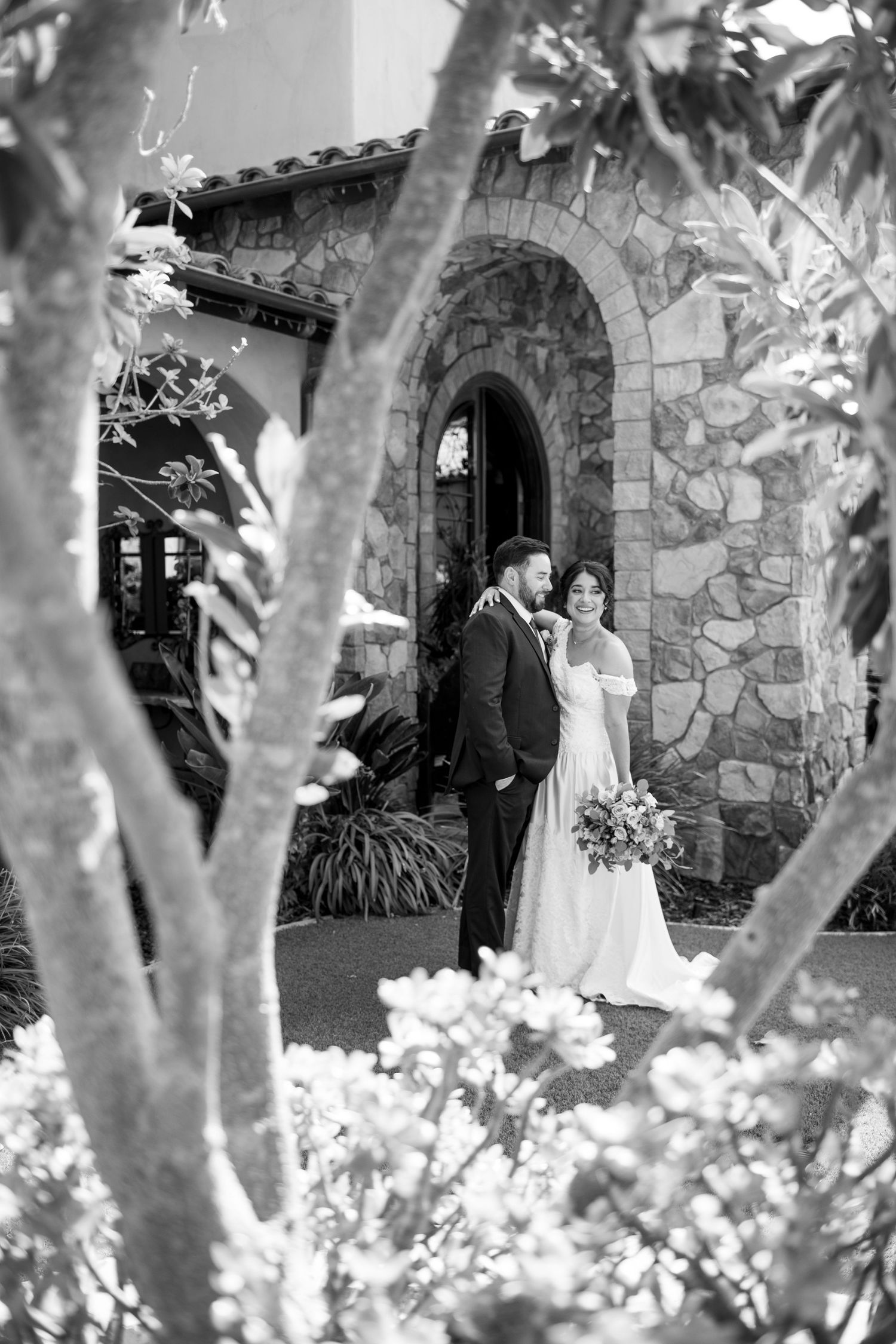 A wedding couple poses in front of a stone archway surrounded by lush garden foliage in black and white.