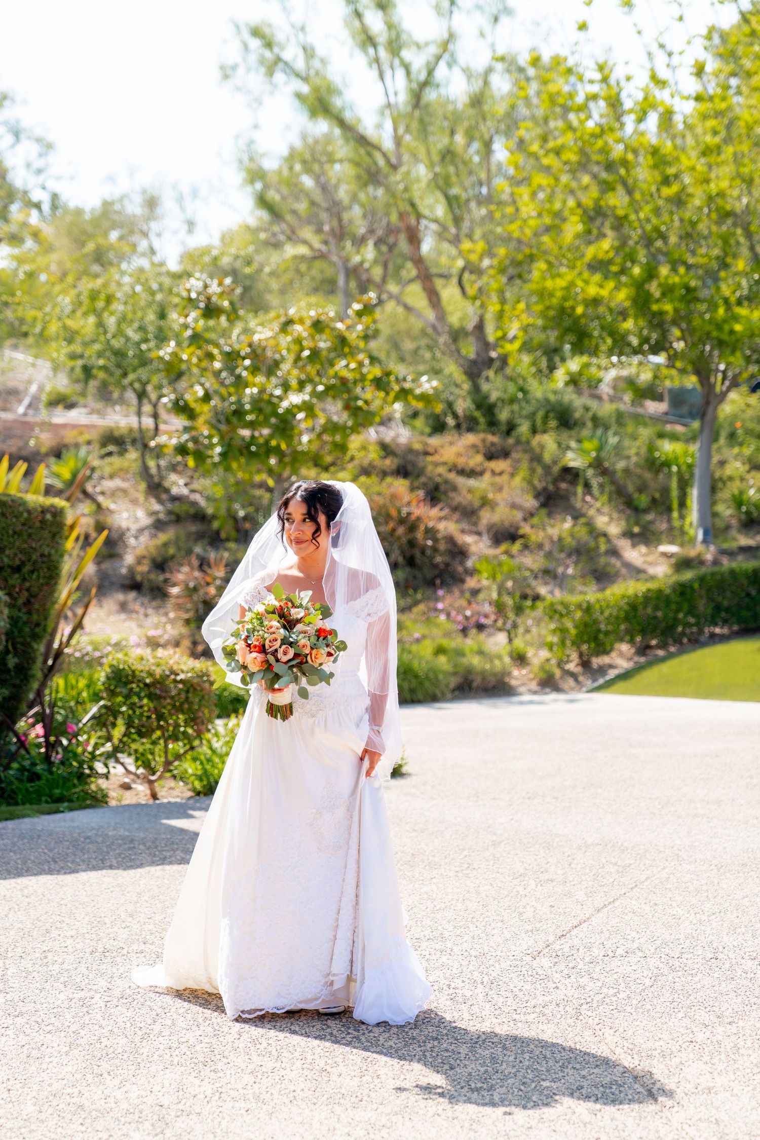 A bride in an elegant white dress stands on a sunlit path holding a vibrant floral bouquet.