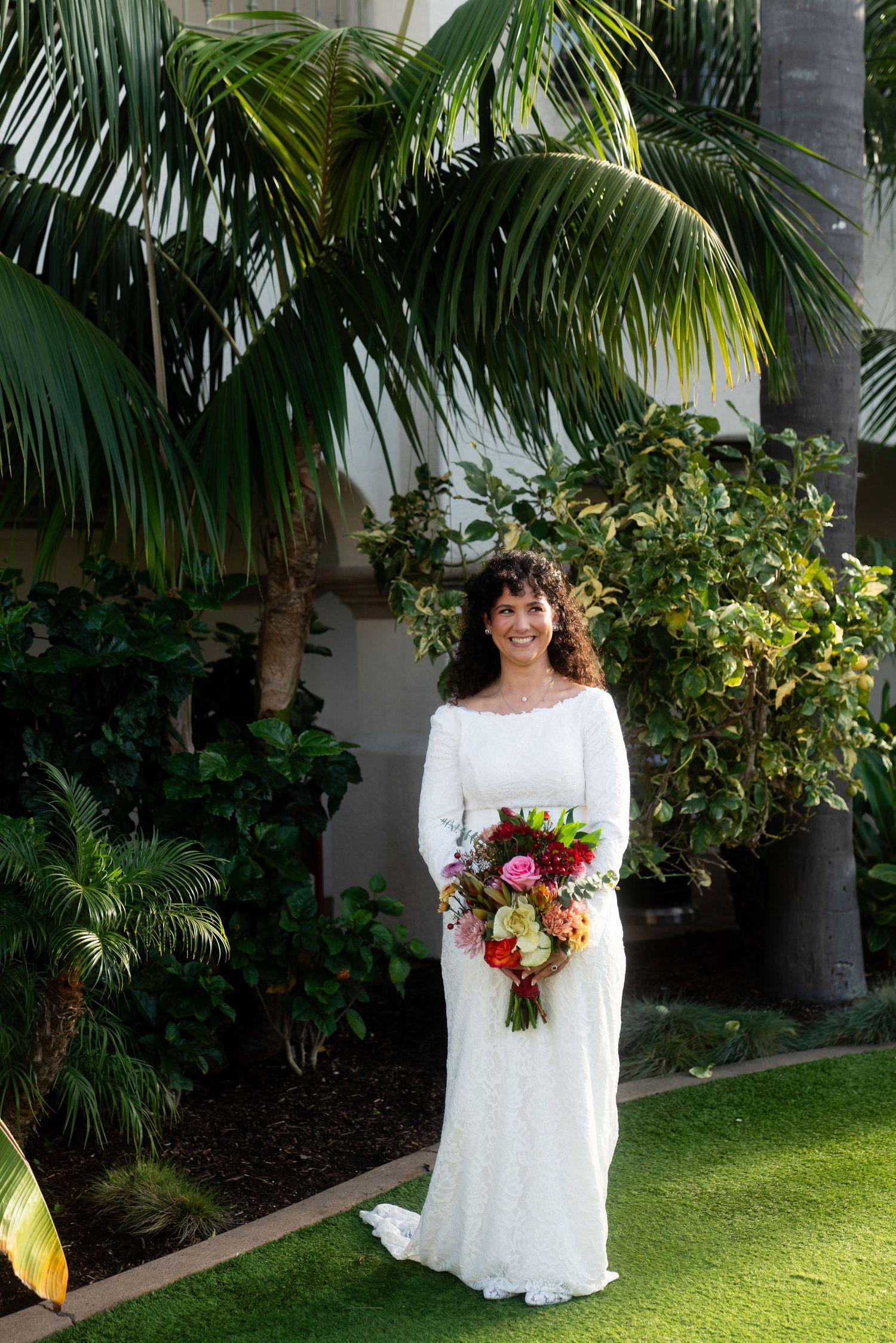 A bride in a long-sleeved white wedding dress stands in a lush tropical garden setting holding a colorful bouquet.