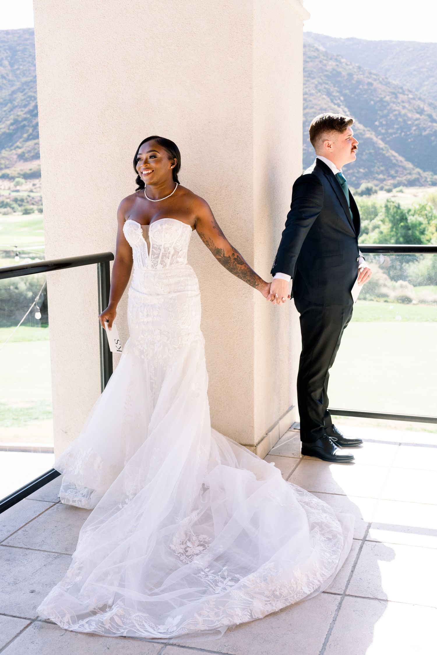 A bride in a stunning white mermaid gown holds hands with someone in a black suit against a mountain backdrop.