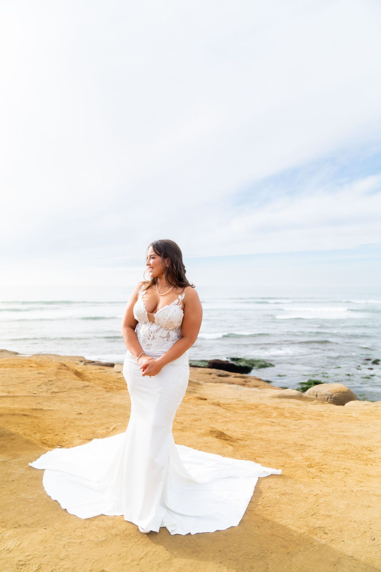A bride in a white fitted dress poses on a cliff overlooking the ocean.