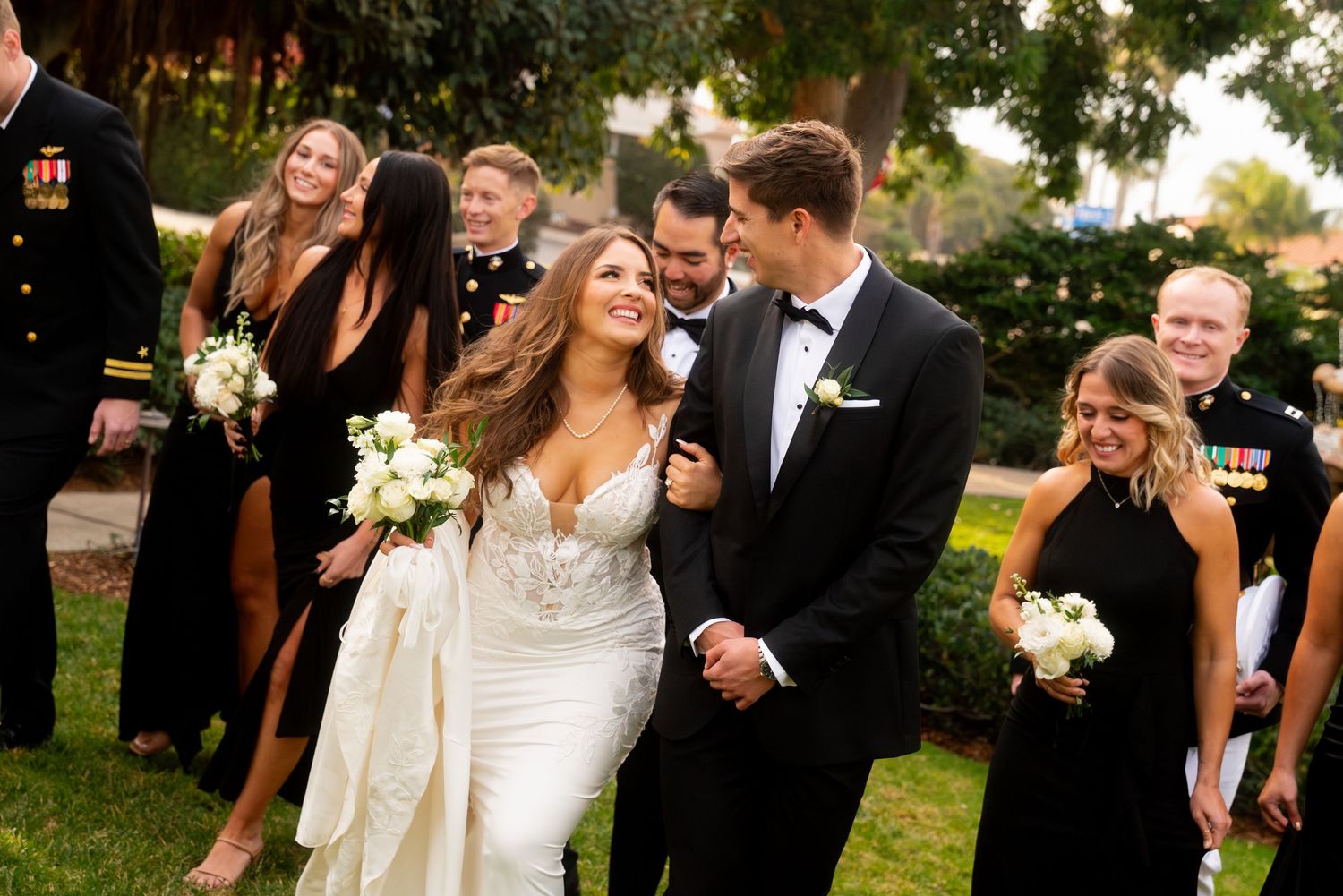 Joyful wedding party in formal attire laughing together outdoors with bride in lace gown and groom in tuxedo.