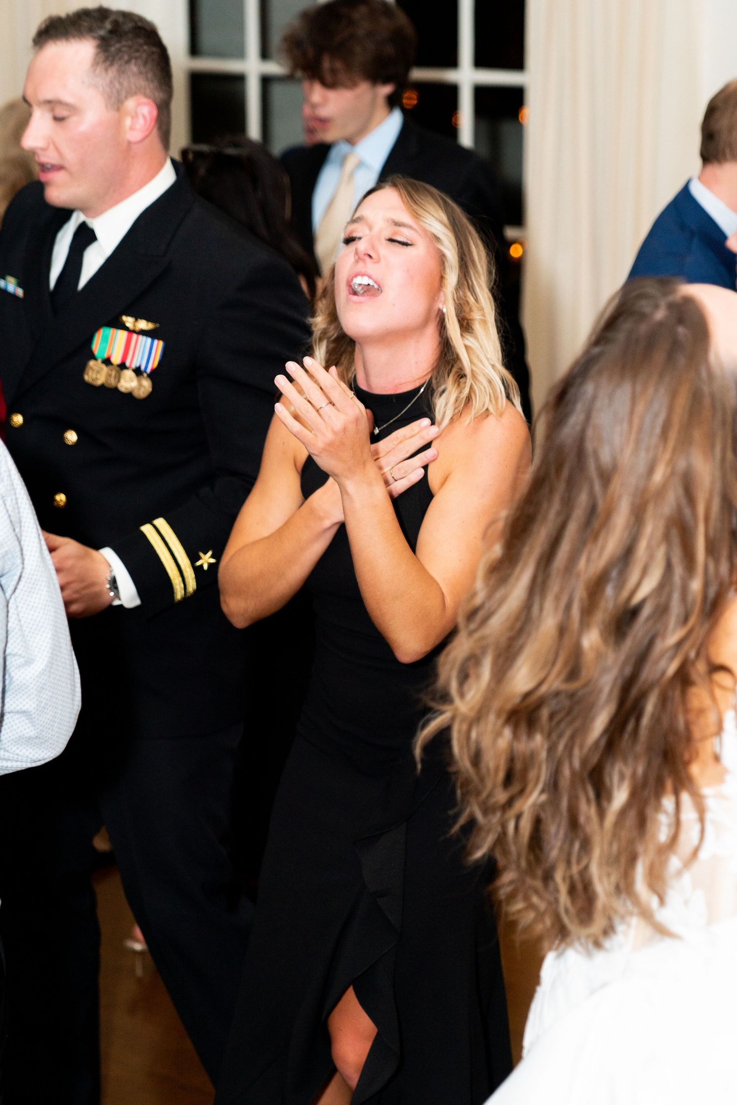 Wedding guest in black dress dancing enthusiastically at reception next to military officer.