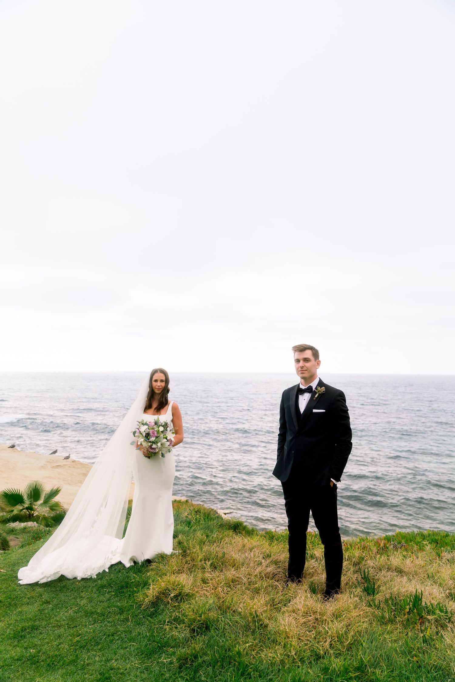 A couple in wedding attire poses on a grassy cliff overlooking the ocean on their special day.