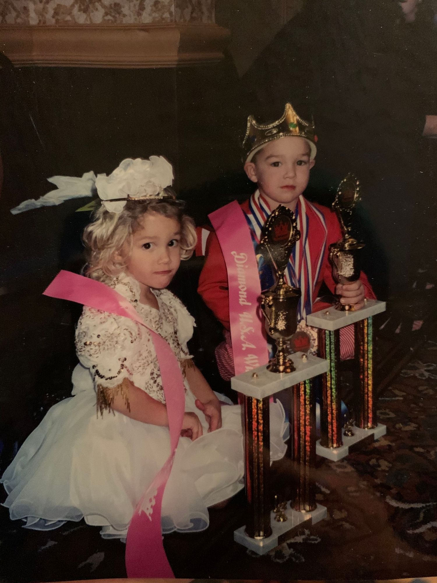Two pageant winners wearing formal attire and sashes stand together displaying their trophies at an event.
