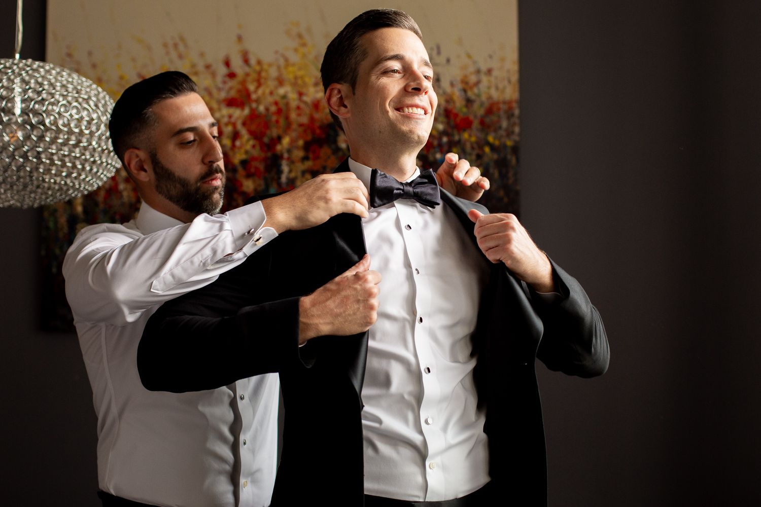 A groomsman helps adjust another man's bowtie and suit jacket while getting ready in a warmly lit room.