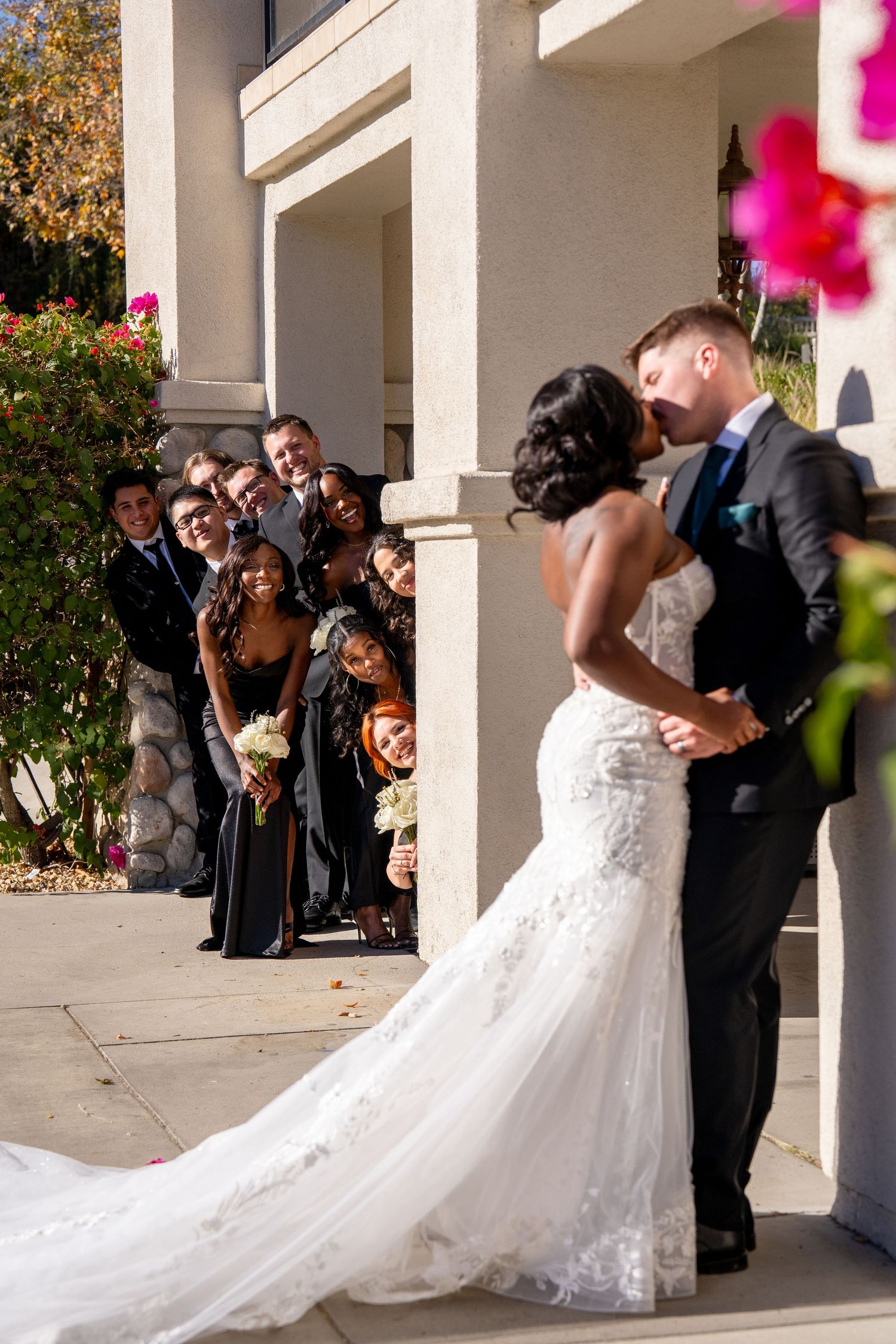 A wedding couple shares a romantic moment against white pillars while wedding party members look on from behind.
