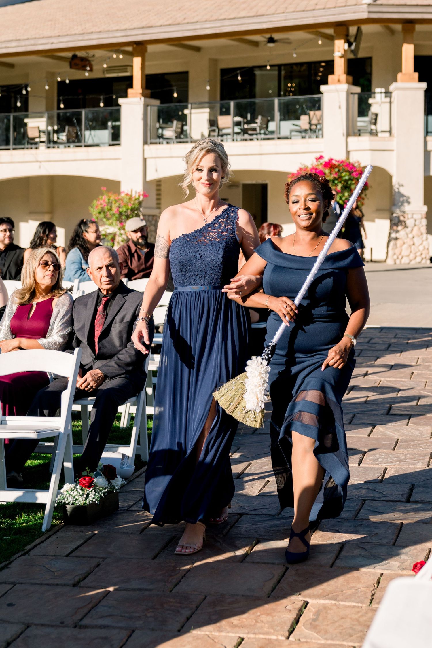 Two guests in elegant blue dresses walk down an aisle at an outdoor wedding ceremony.