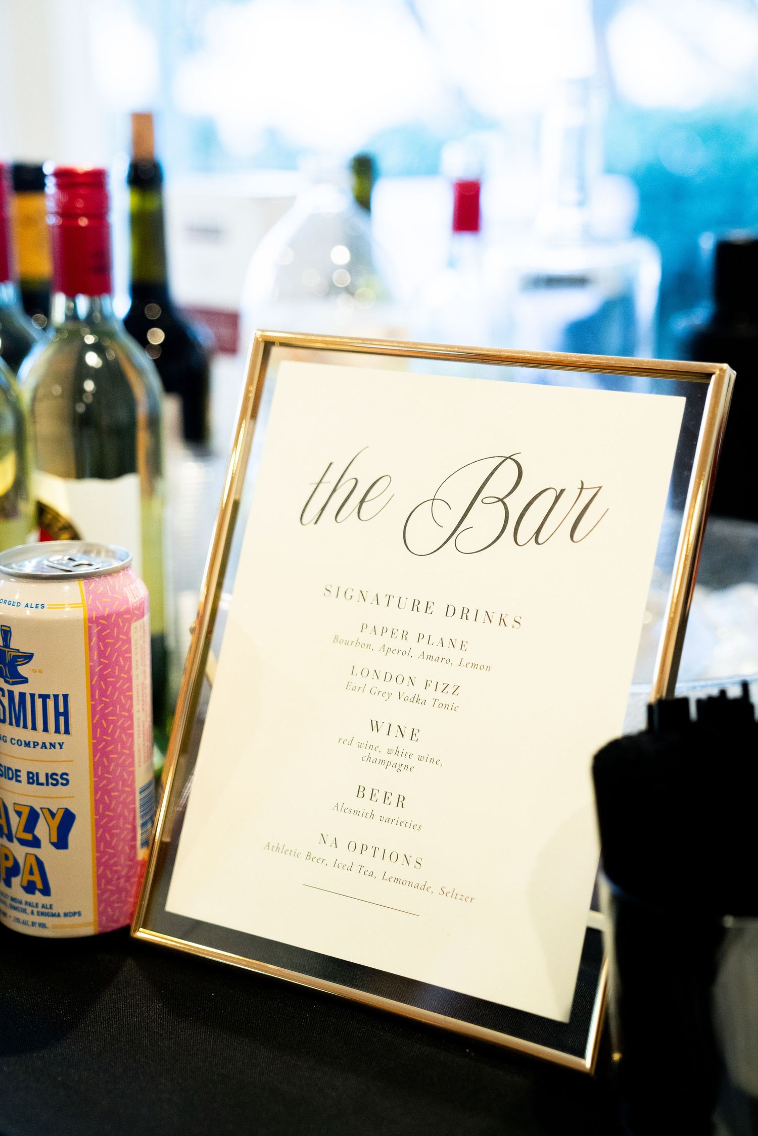 An elegant framed bar menu sign displayed next to wine bottles and craft beer at a wedding reception.