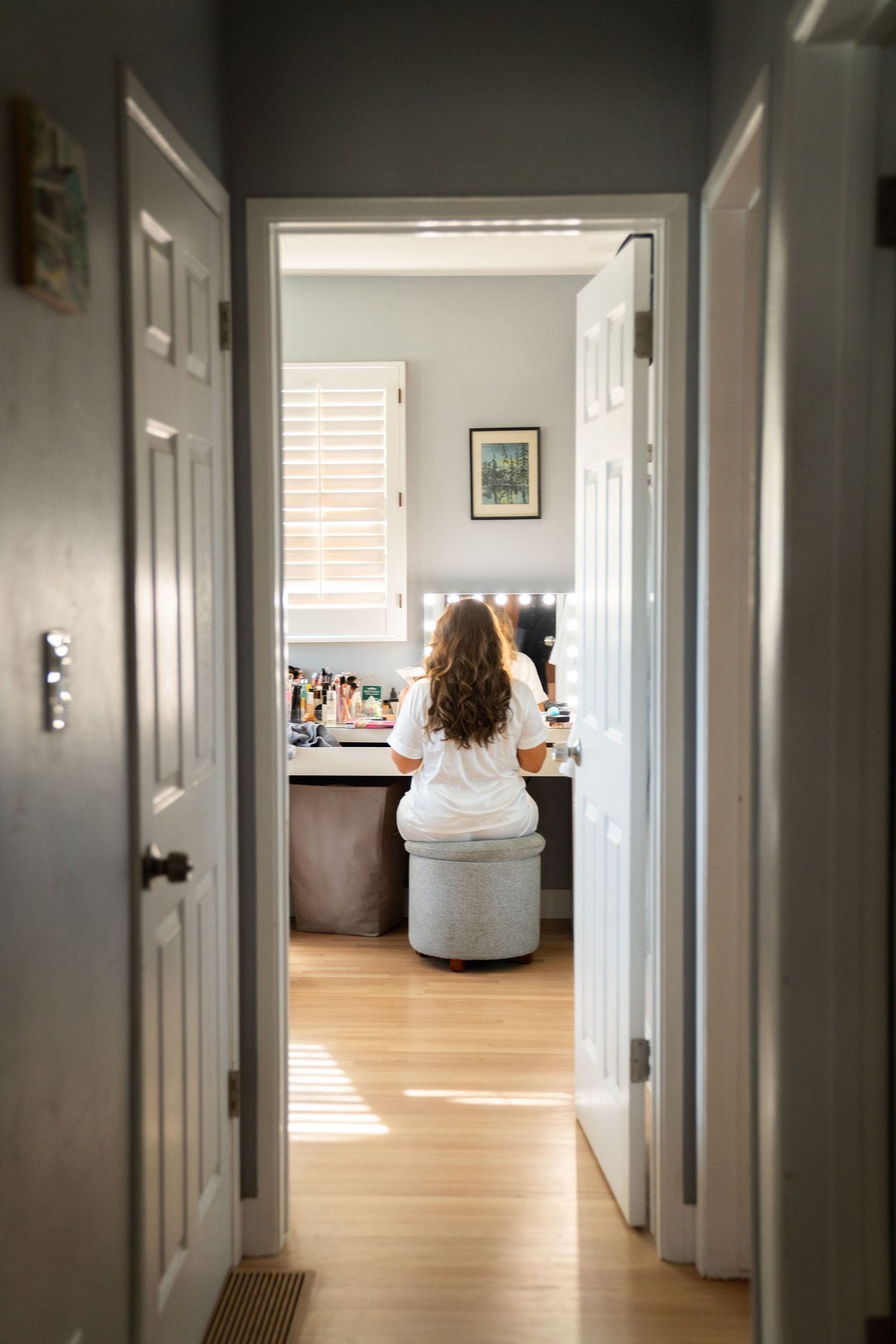View through doorway of someone sitting at a vanity desk in a sunlit room with wooden floors.