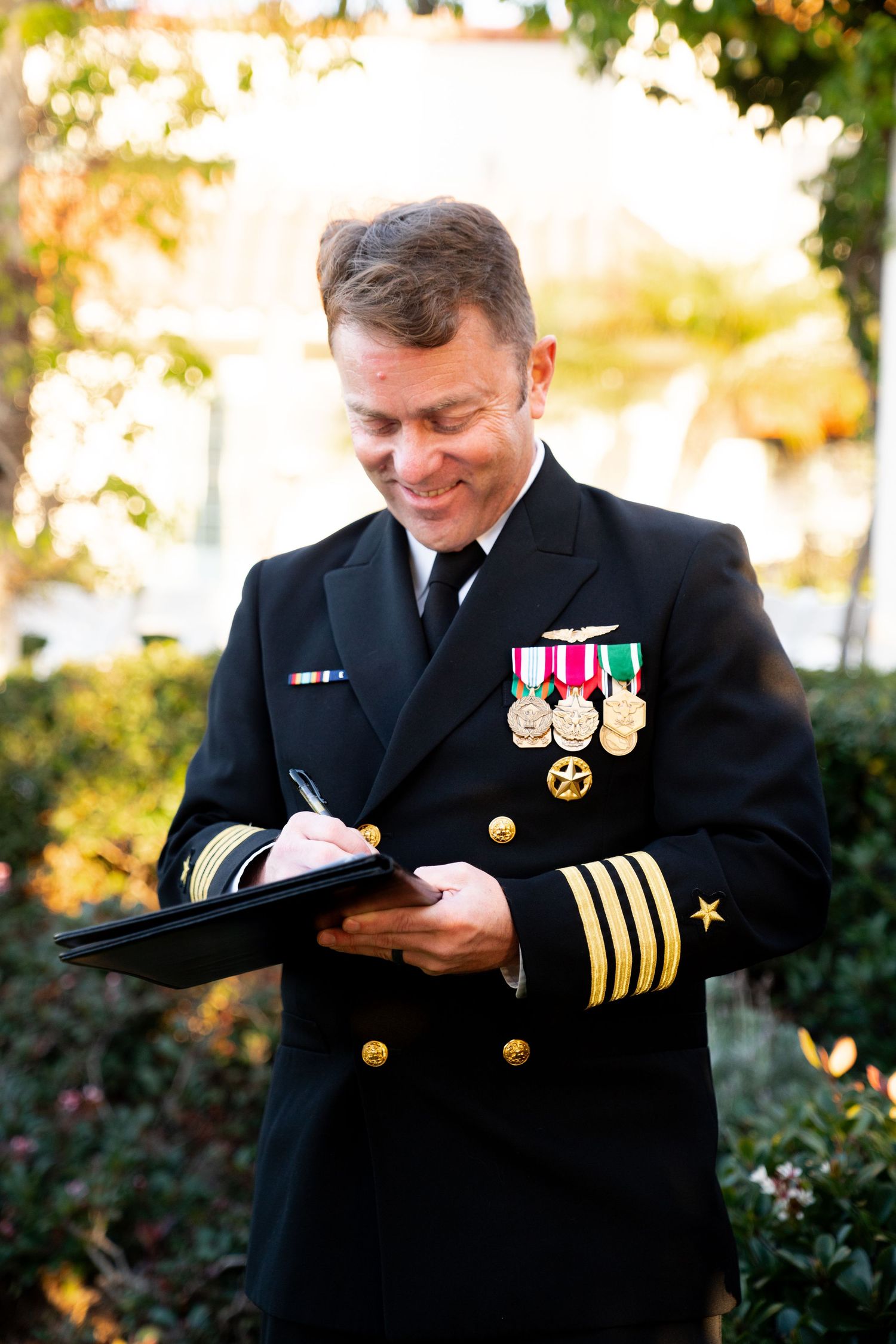 Naval officer in dress uniform with medals writing on clipboard in garden setting.