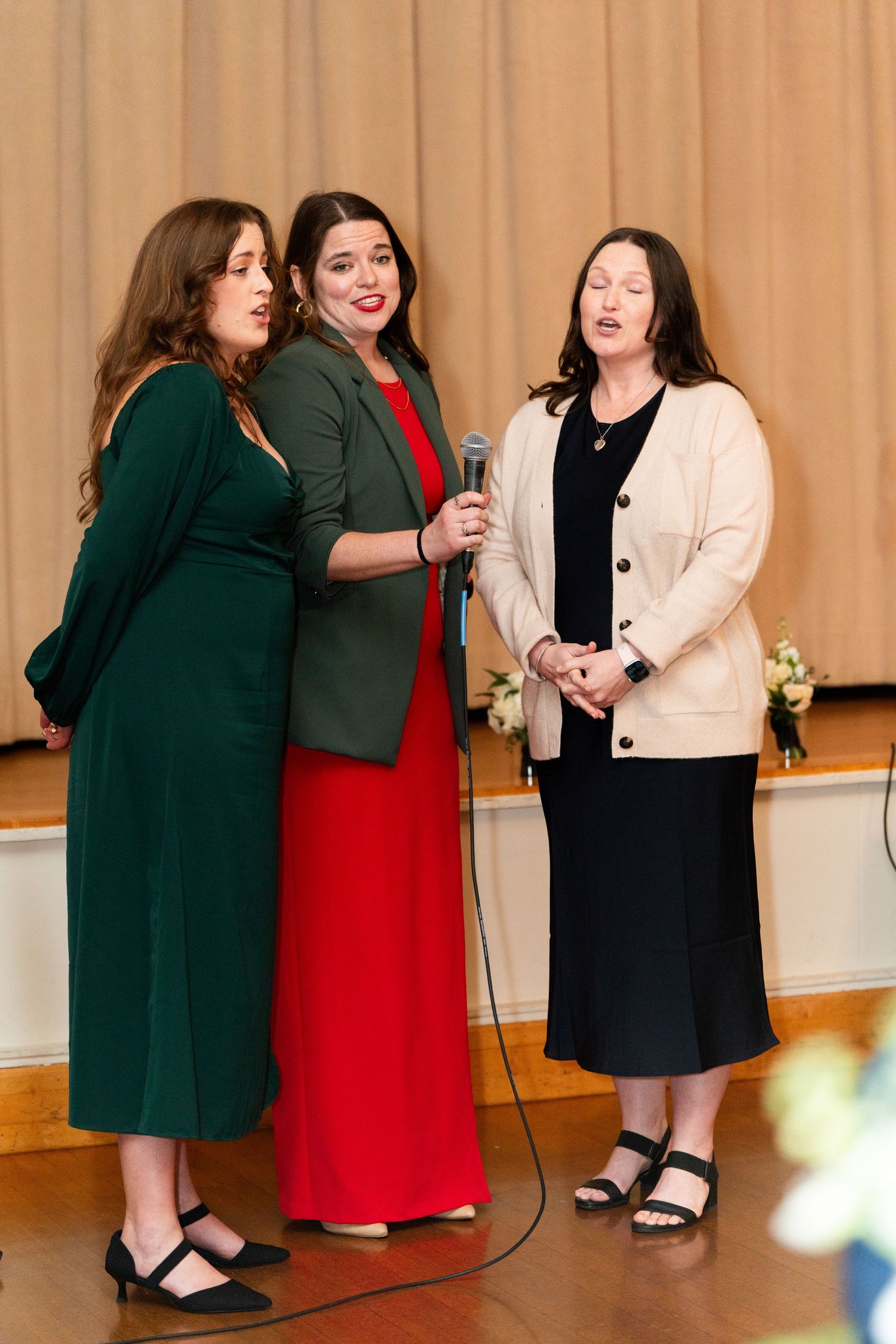 Three women in formal dresses sharing a microphone at an event.