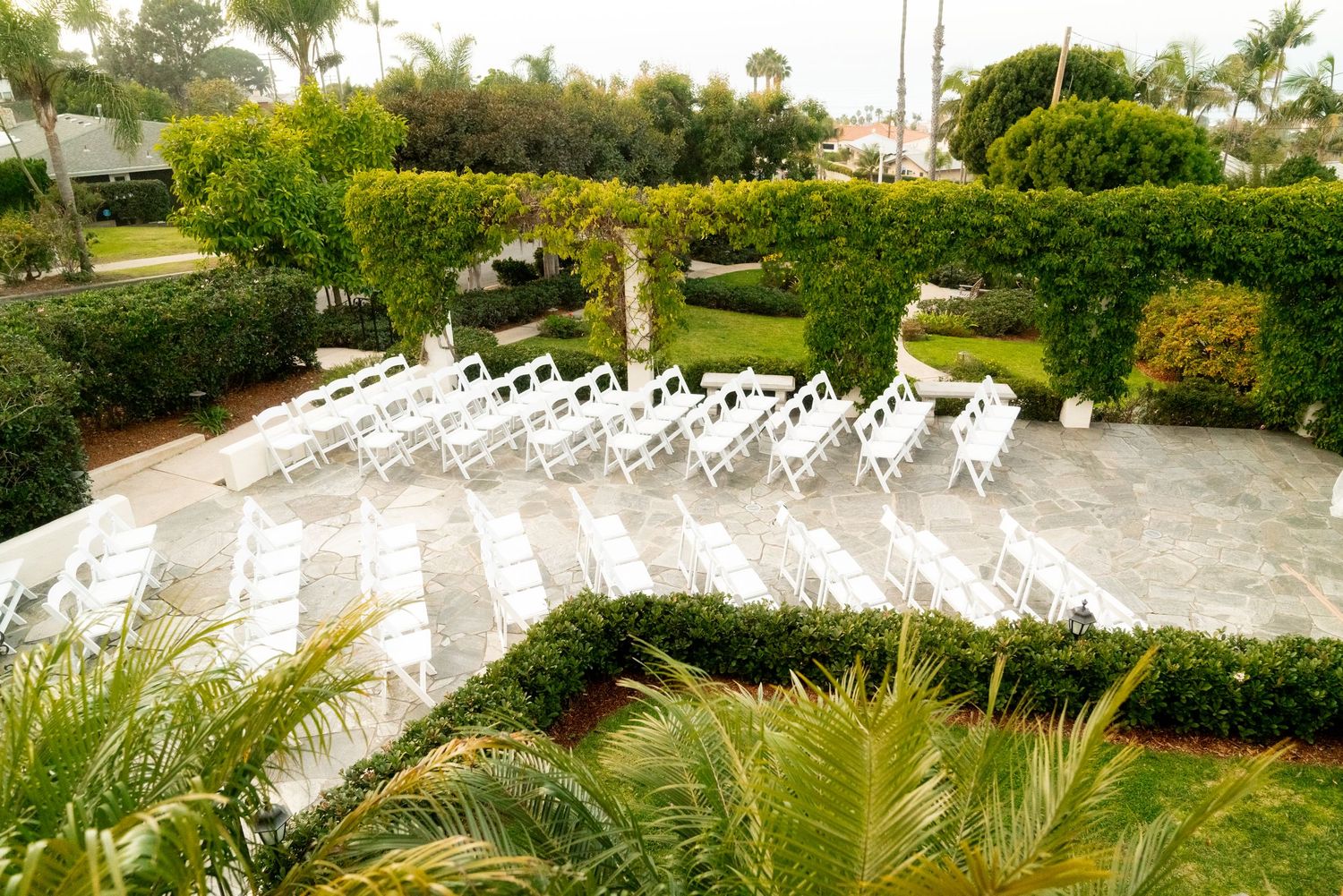 Aerial view of outdoor wedding ceremony setup with white chairs arranged on stone patio surrounded by greenery.