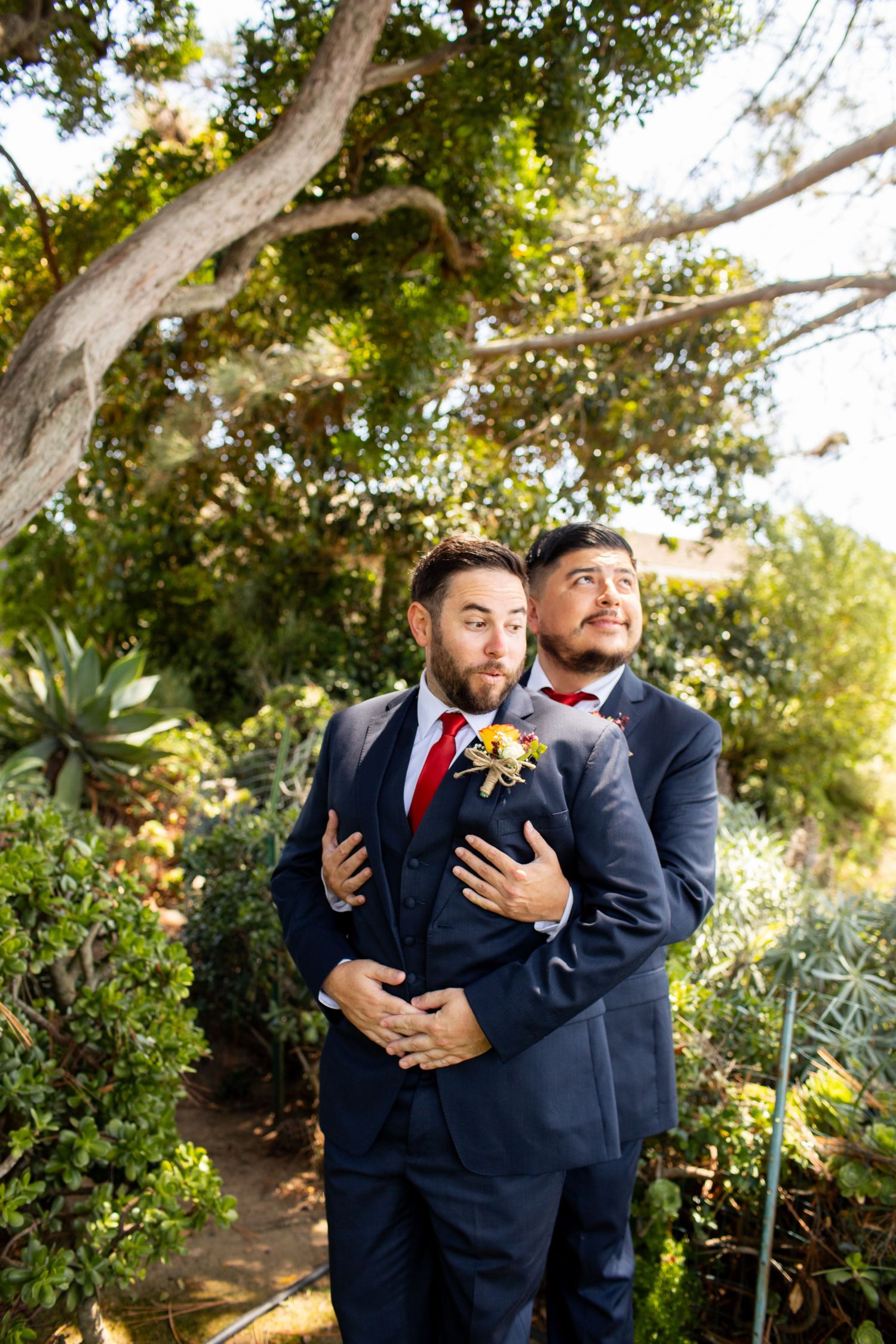 Two people in navy suits share an intimate embrace during wedding photos in a lush garden.
