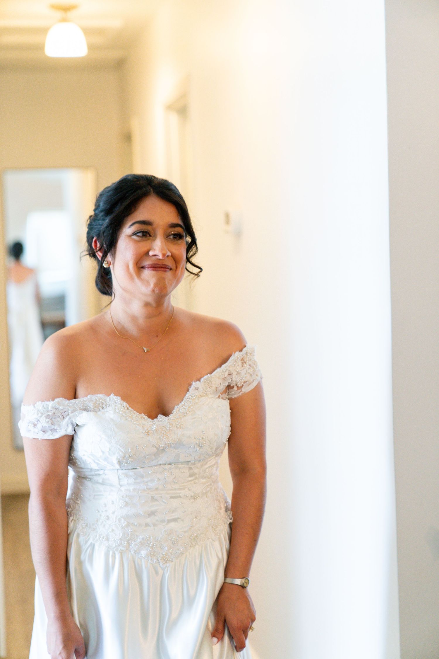 A bride in an elegant off-shoulder lace wedding dress smiles warmly while standing in a bright hallway.