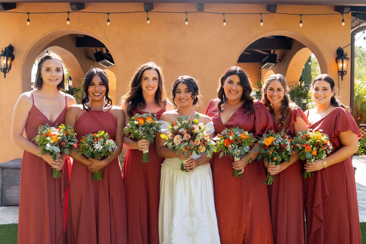 Wedding party poses in terracotta-colored dresses with vibrant bouquets against a Spanish-style backdrop.