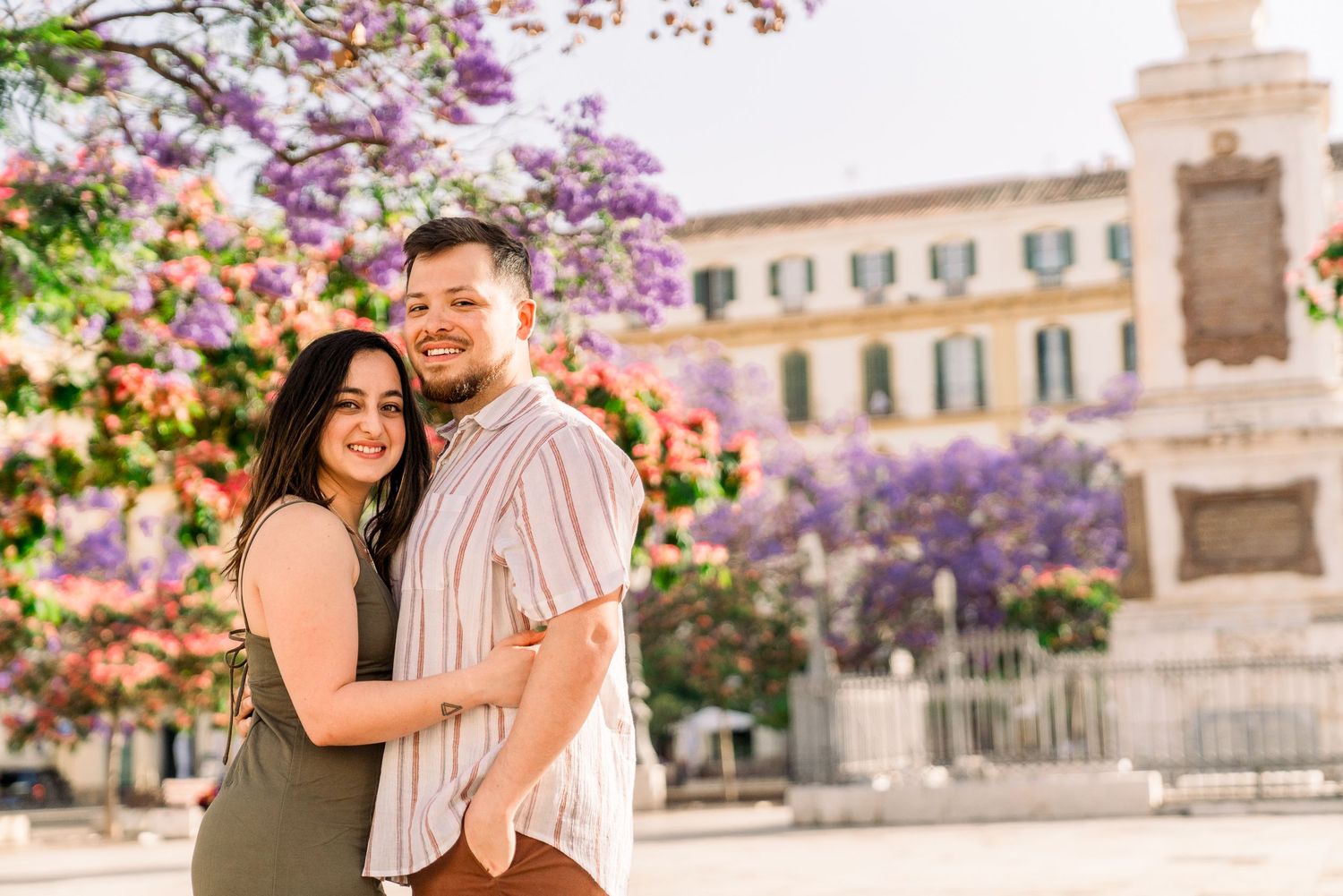 A couple embraces under blooming purple jacaranda trees in a European city plaza.