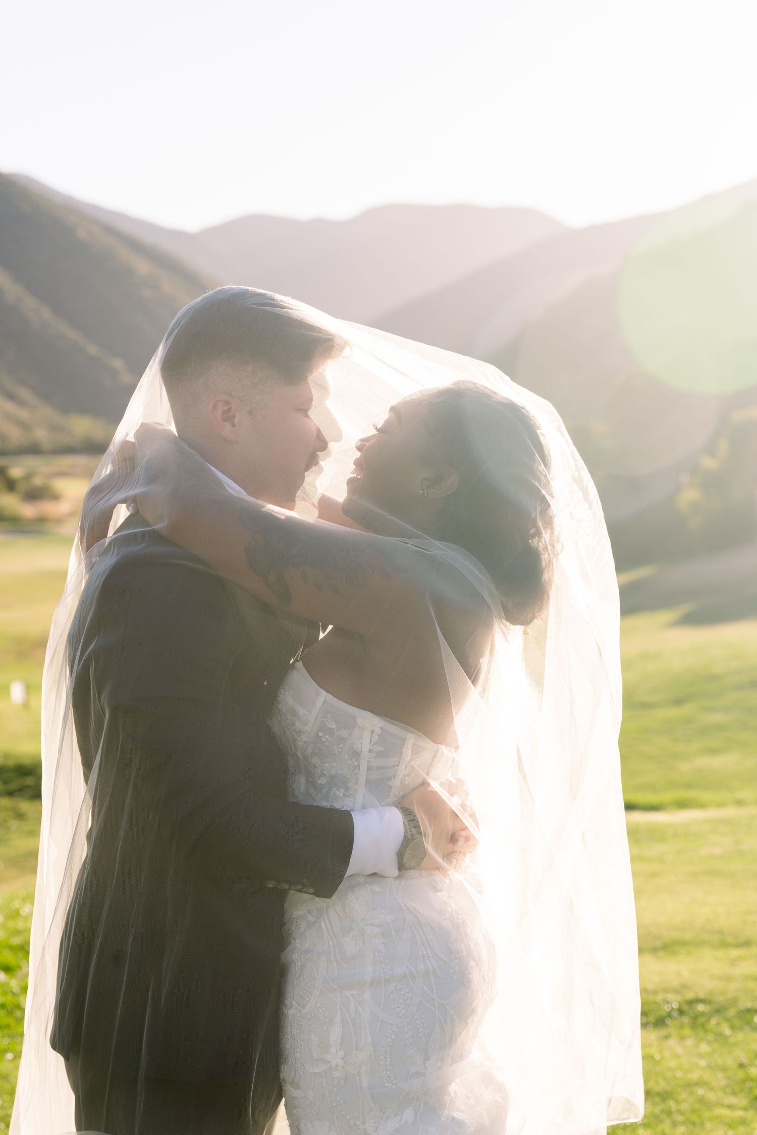 A romantic wedding couple embraces under a flowing veil with sunlit mountains in the background.