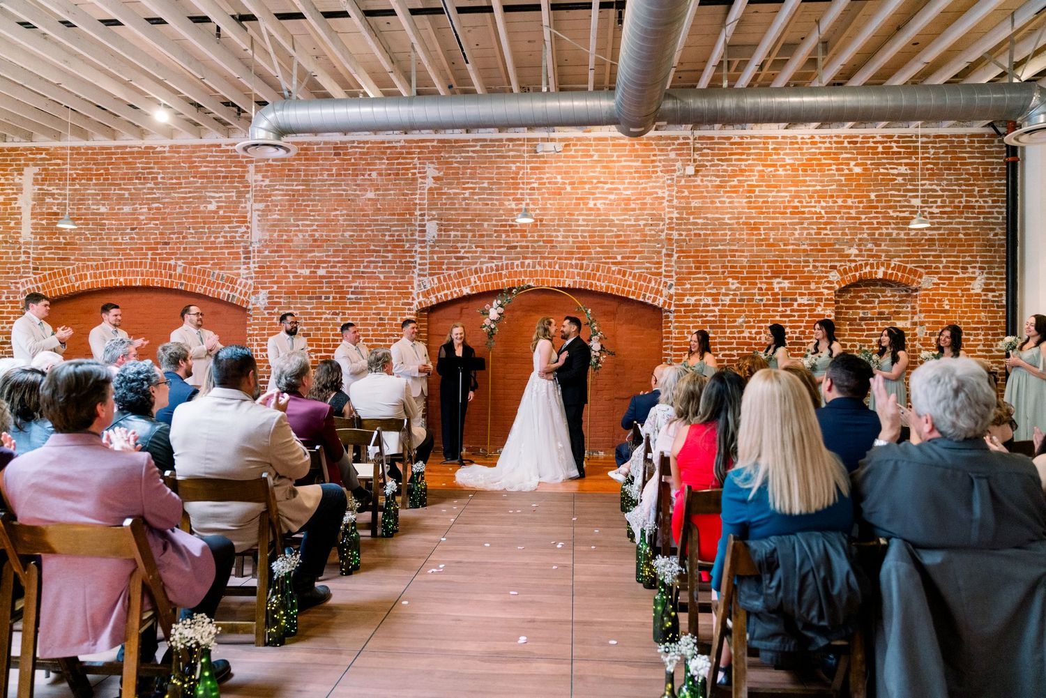 Indoor wedding ceremony in an industrial brick venue with exposed ceiling and guests seated in wooden chairs.