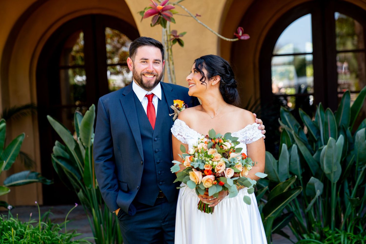 Newlywed couple poses with colorful bouquet against Spanish-style architecture and tropical plants.