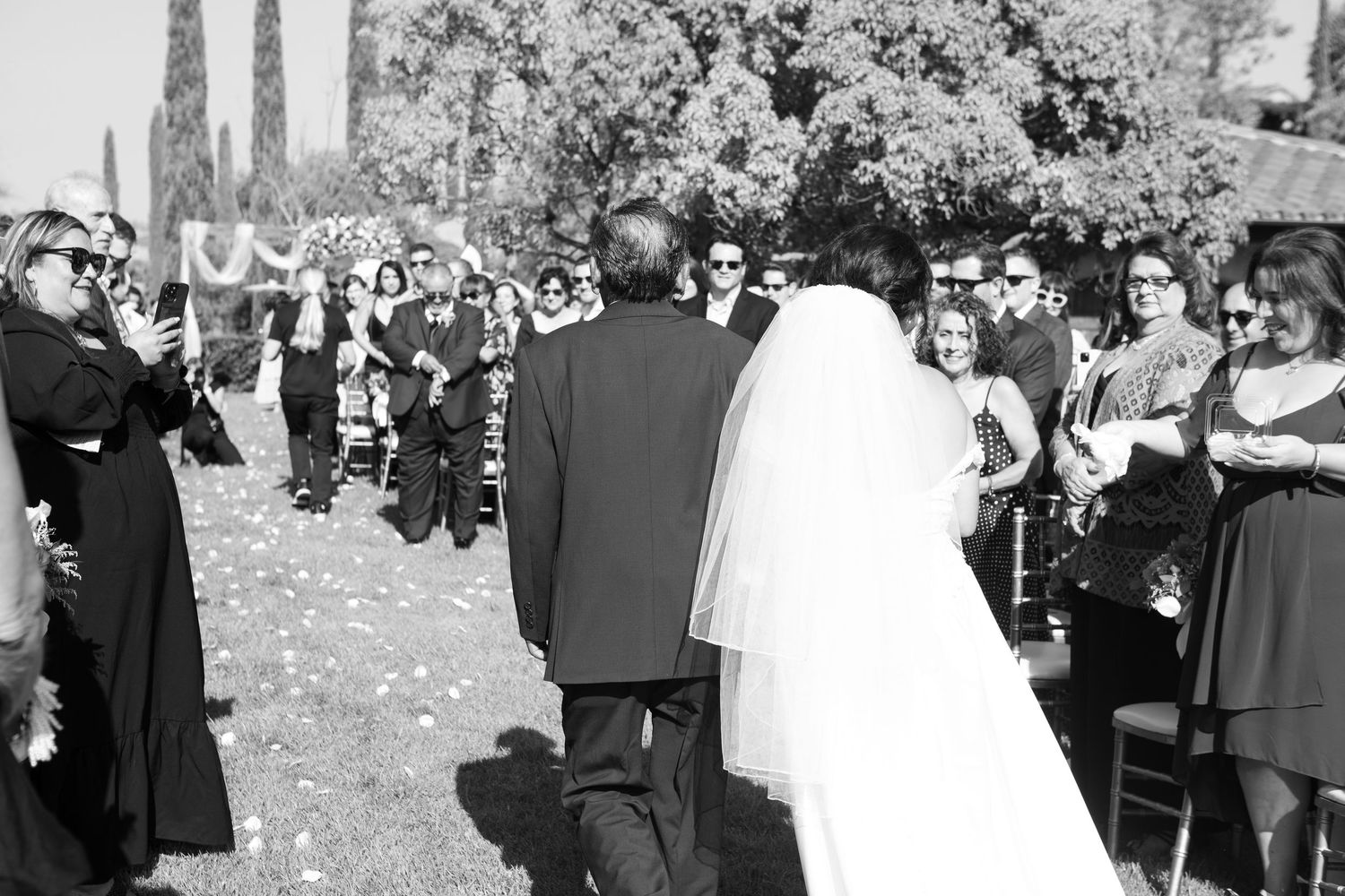 A black and white photo of a wedding ceremony with guests watching the couple from rows of chairs outdoors.