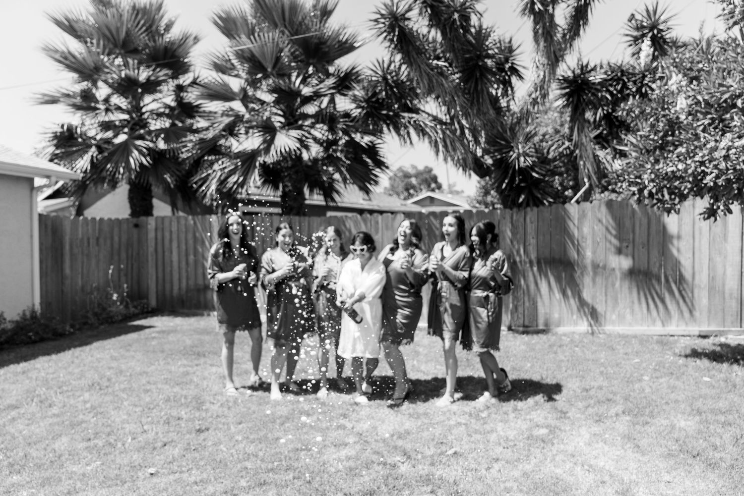 A black and white photograph shows a group of people gathered outdoors in front of palm trees on a sunny day.