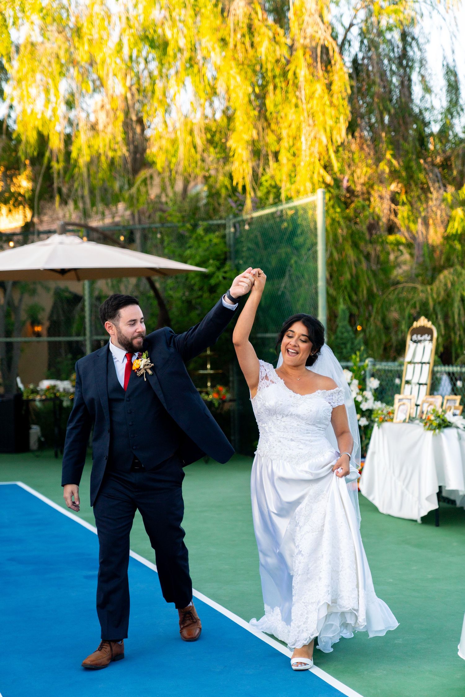 A joyful couple dances together on a blue floor at an outdoor wedding reception with yellow willow trees in background.