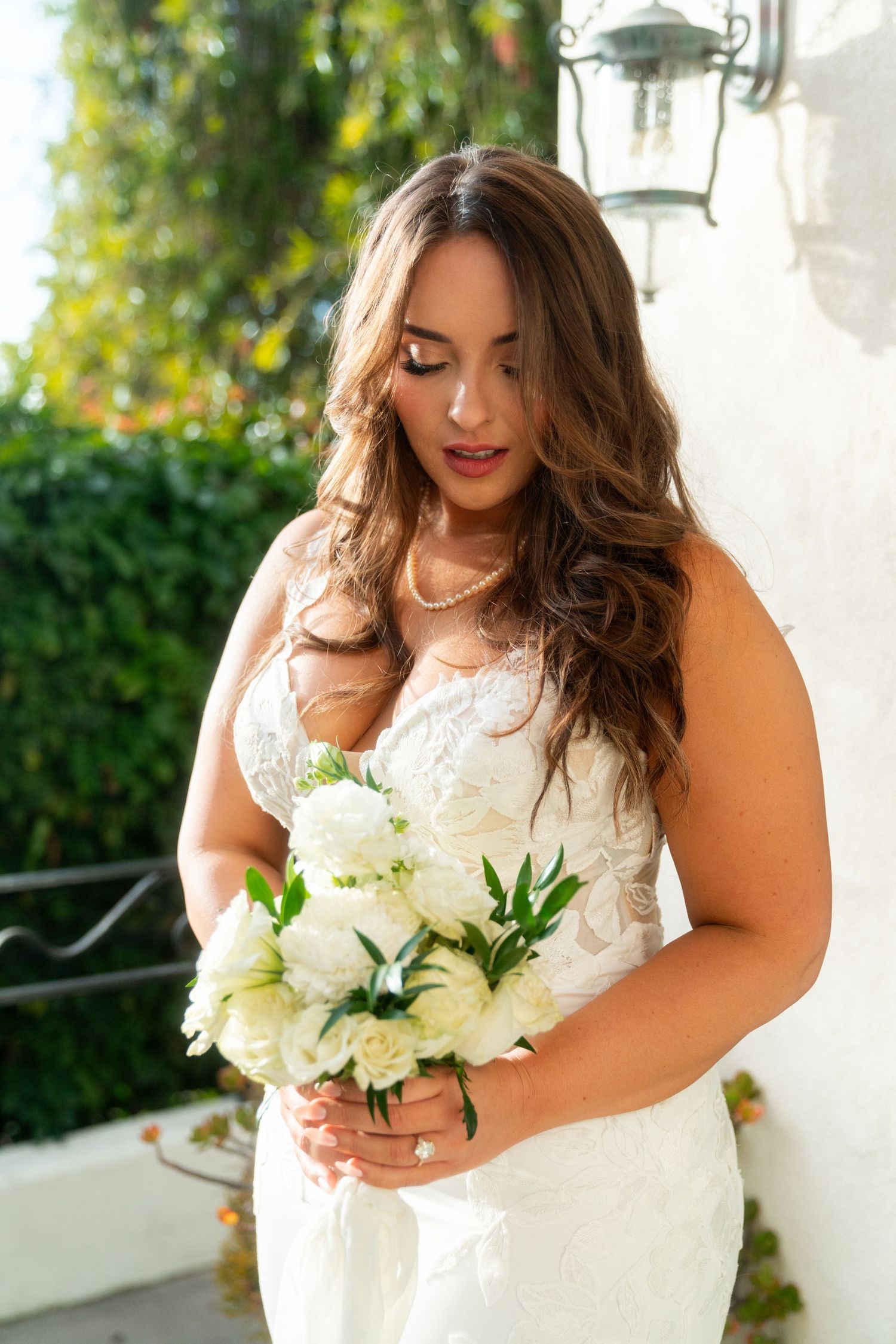 A bride in a white lace dress holds a bouquet of white roses outdoors while looking down with red lipstick.