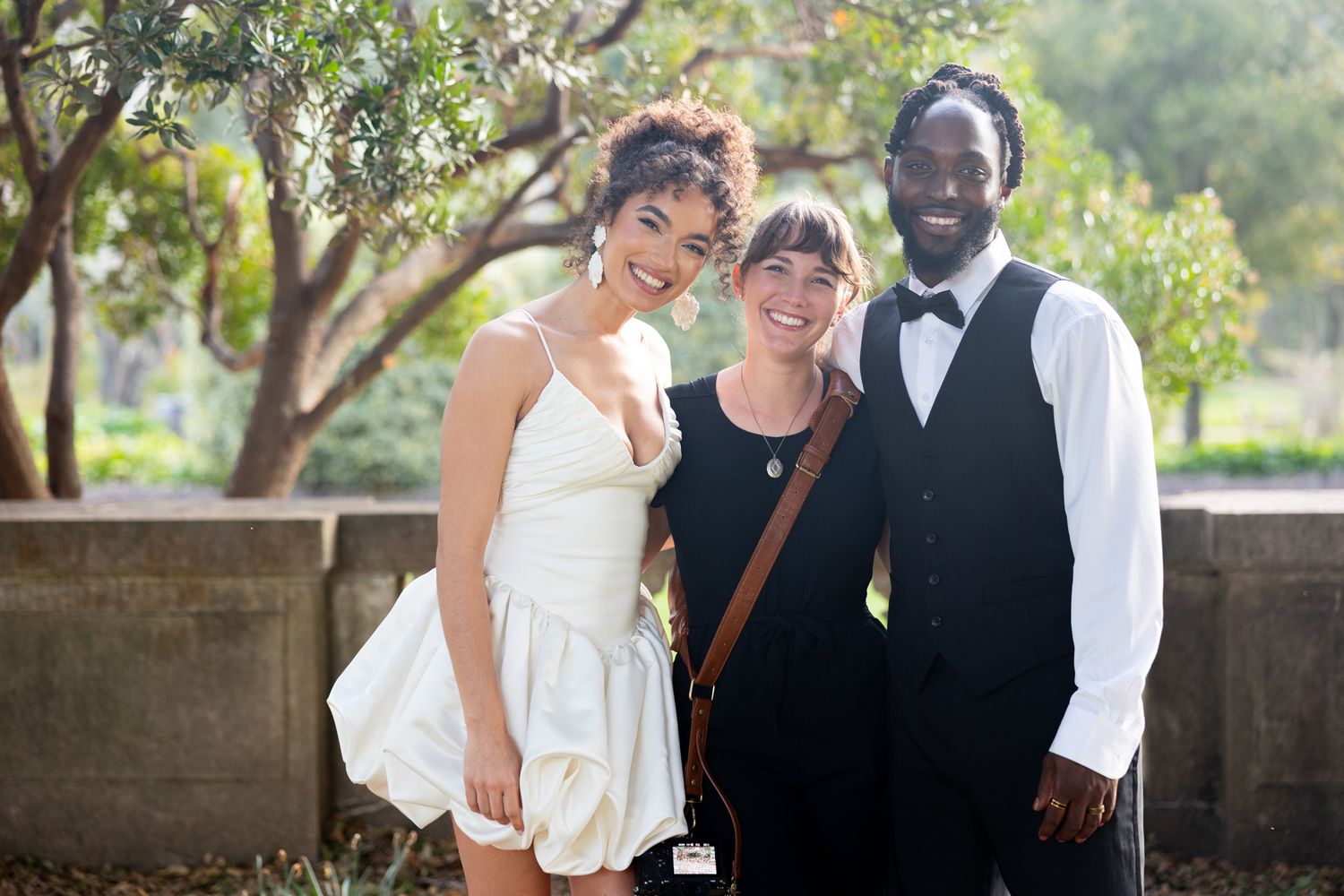 Three people pose together outdoors in formal attire at a garden event with trees in the background.