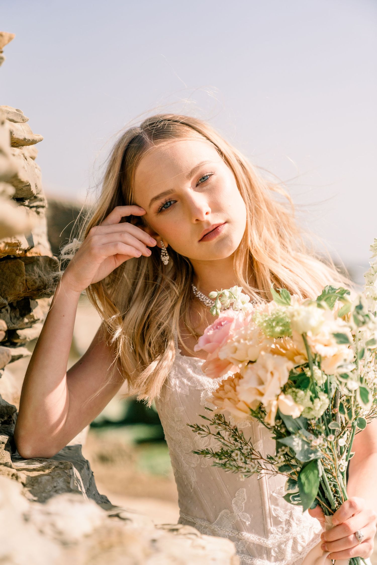 Portrait of person in white lace dress holding delicate floral bouquet against sunlit stone backdrop.