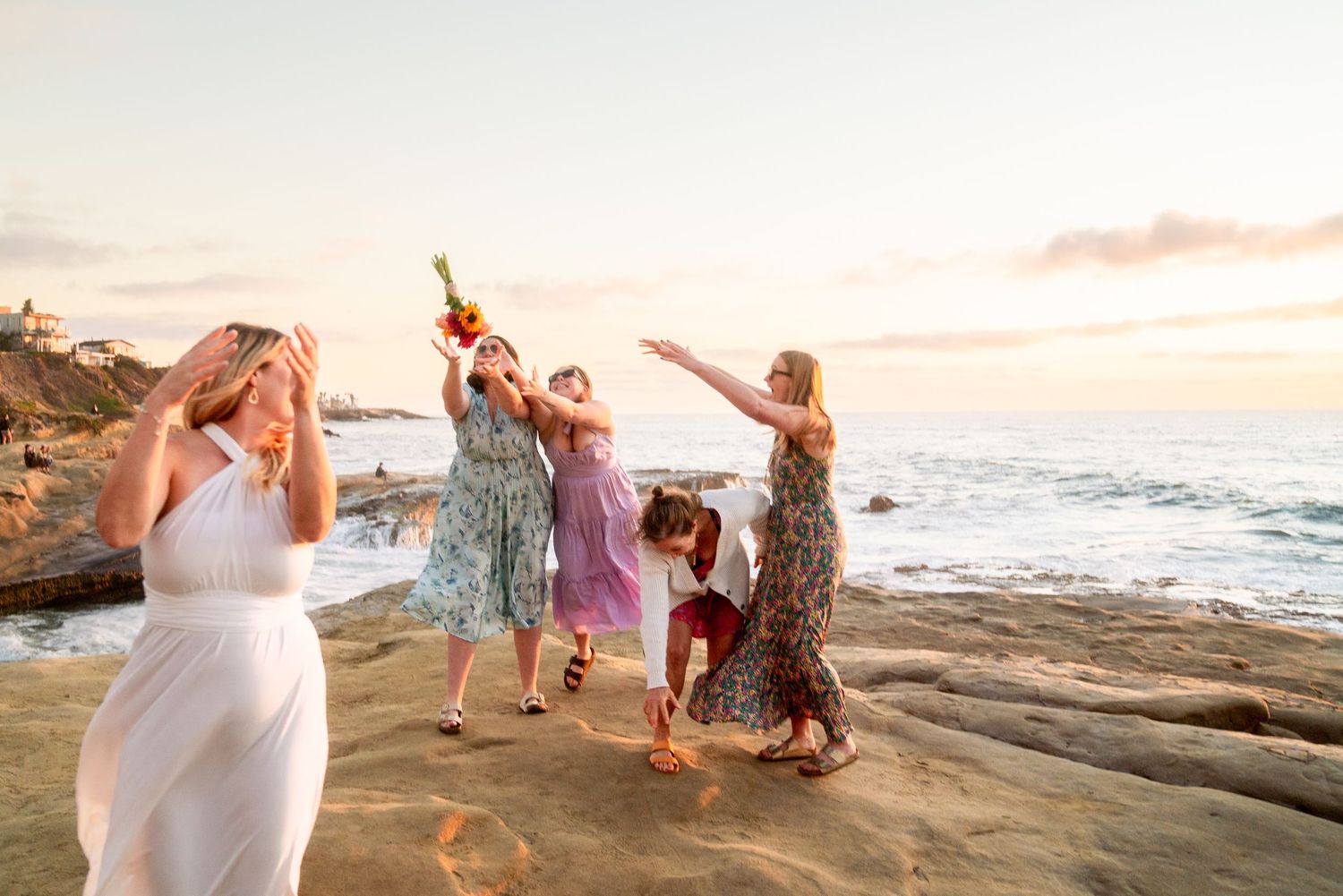 A group of women dancing joyfully on coastal rocks at sunset by the ocean.
