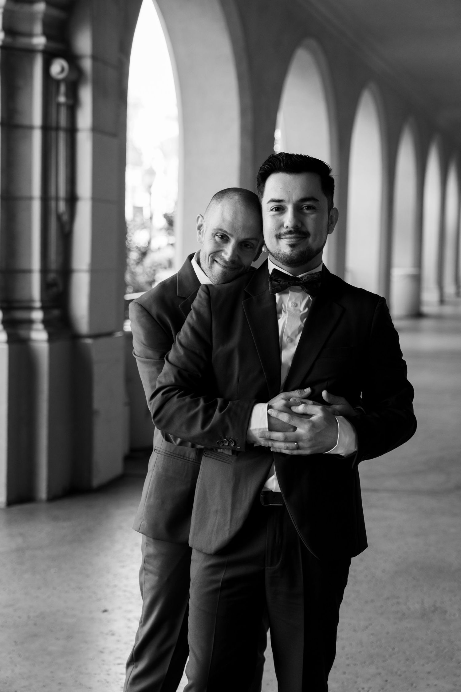 Two people in formal attire share a candid moment under arched columns in black and white photography.