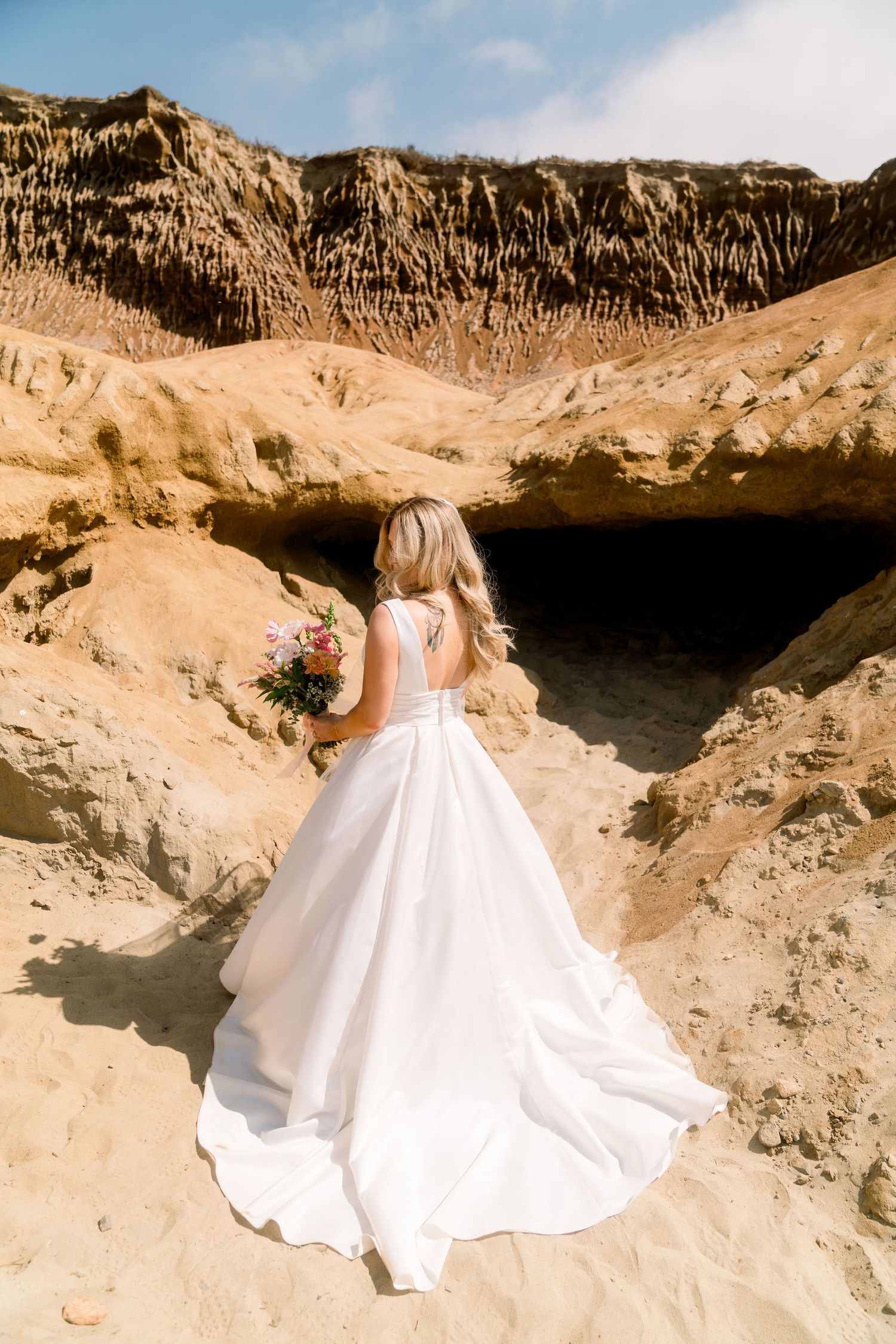 A person in a flowing white wedding dress stands on sandy cliffs holding a bouquet against a dramatic desert landscape.