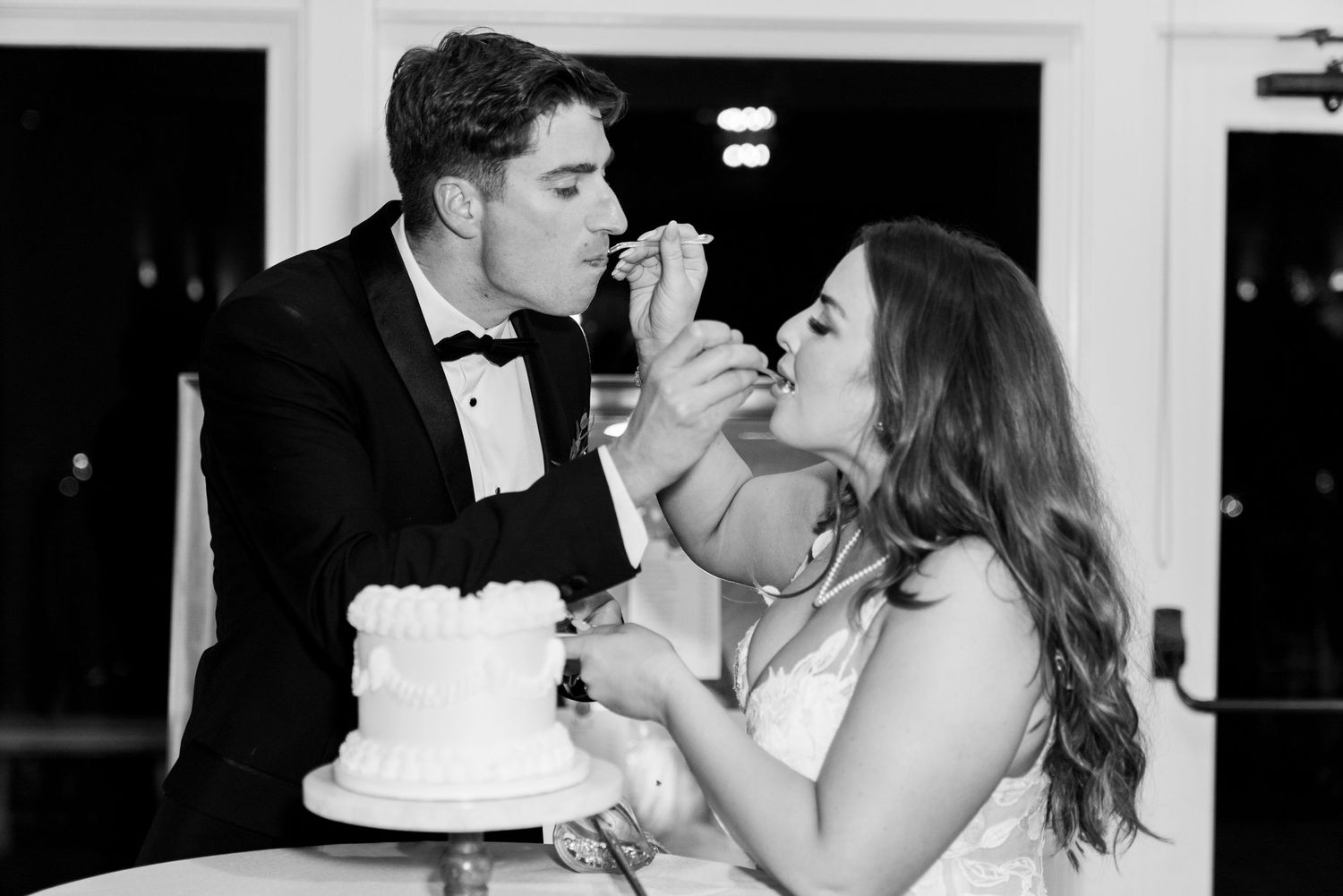 Black and white photo captures playful moment of couple feeding each other wedding cake.