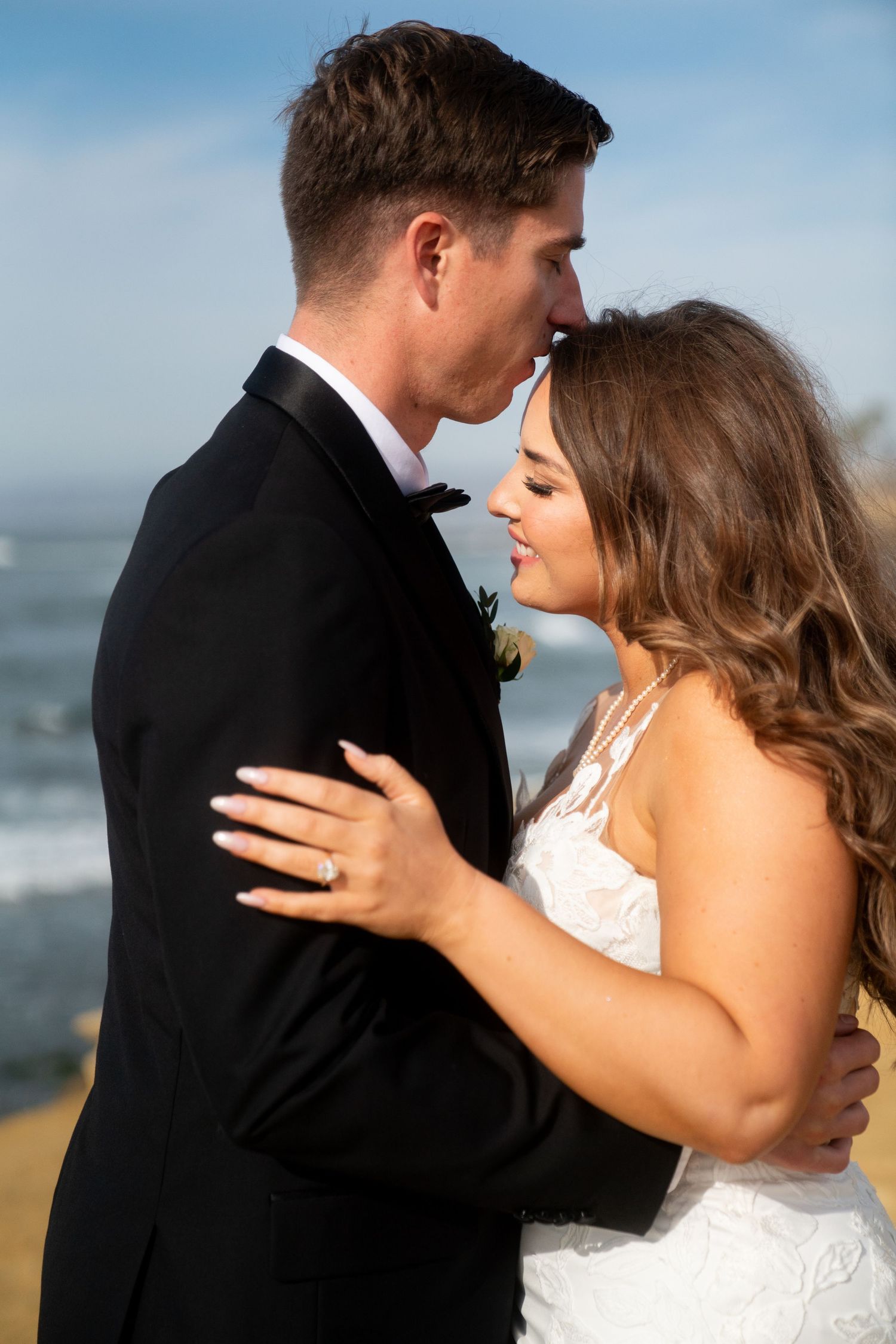 Intimate wedding portrait shows couple embracing at beach with ocean and blue sky in background.