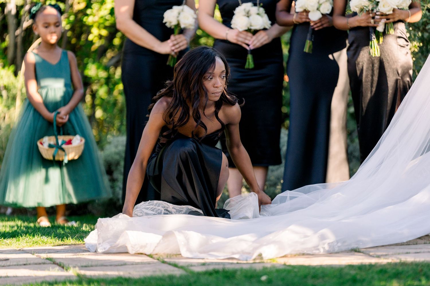 Bridesmaid in black dress arranging bride's long white train during outdoor wedding.