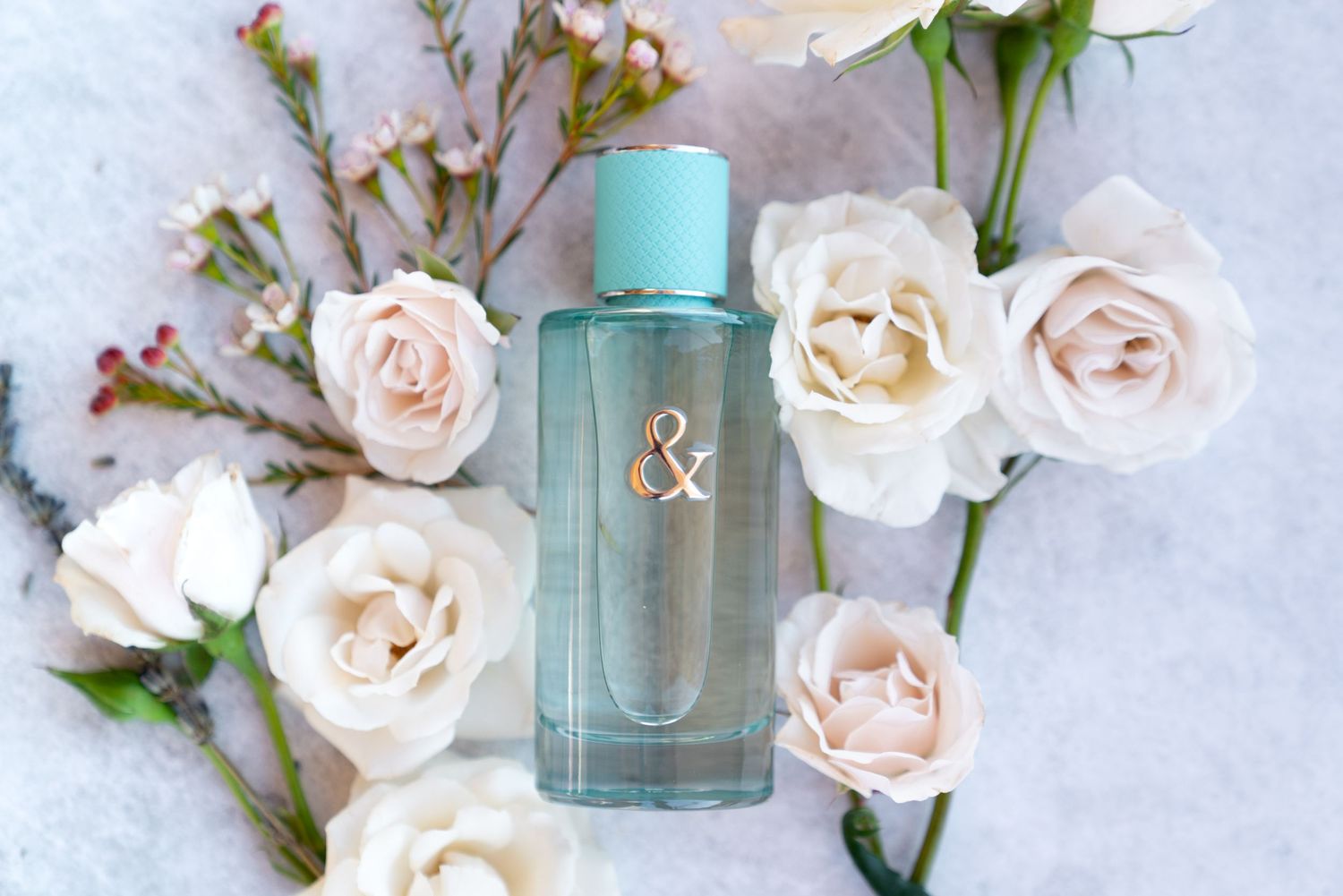 Elegant turquoise perfume bottle surrounded by delicate pink and cream roses on light background.