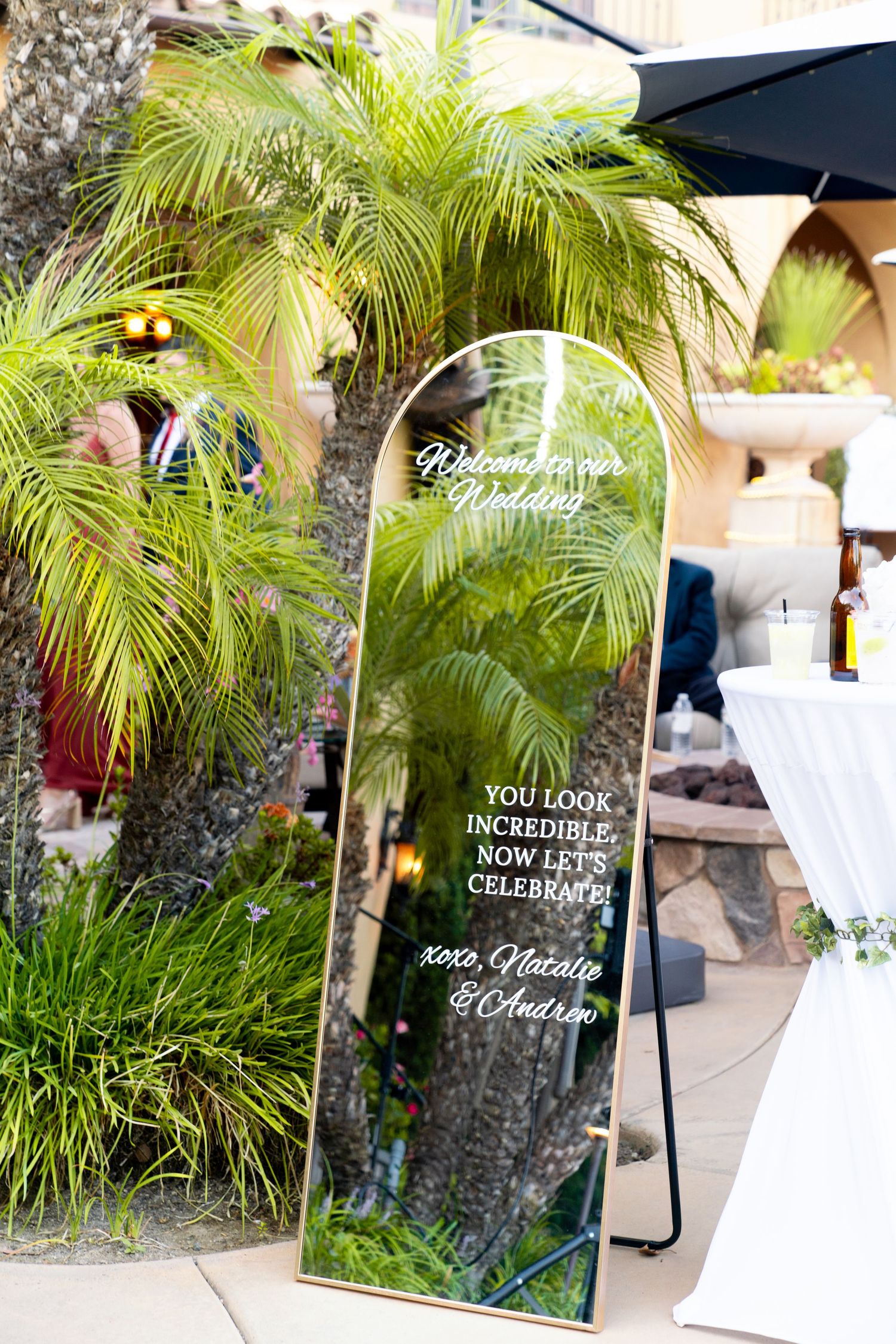Decorative wedding mirror sign with palm trees reflection stands in tropical outdoor setting.