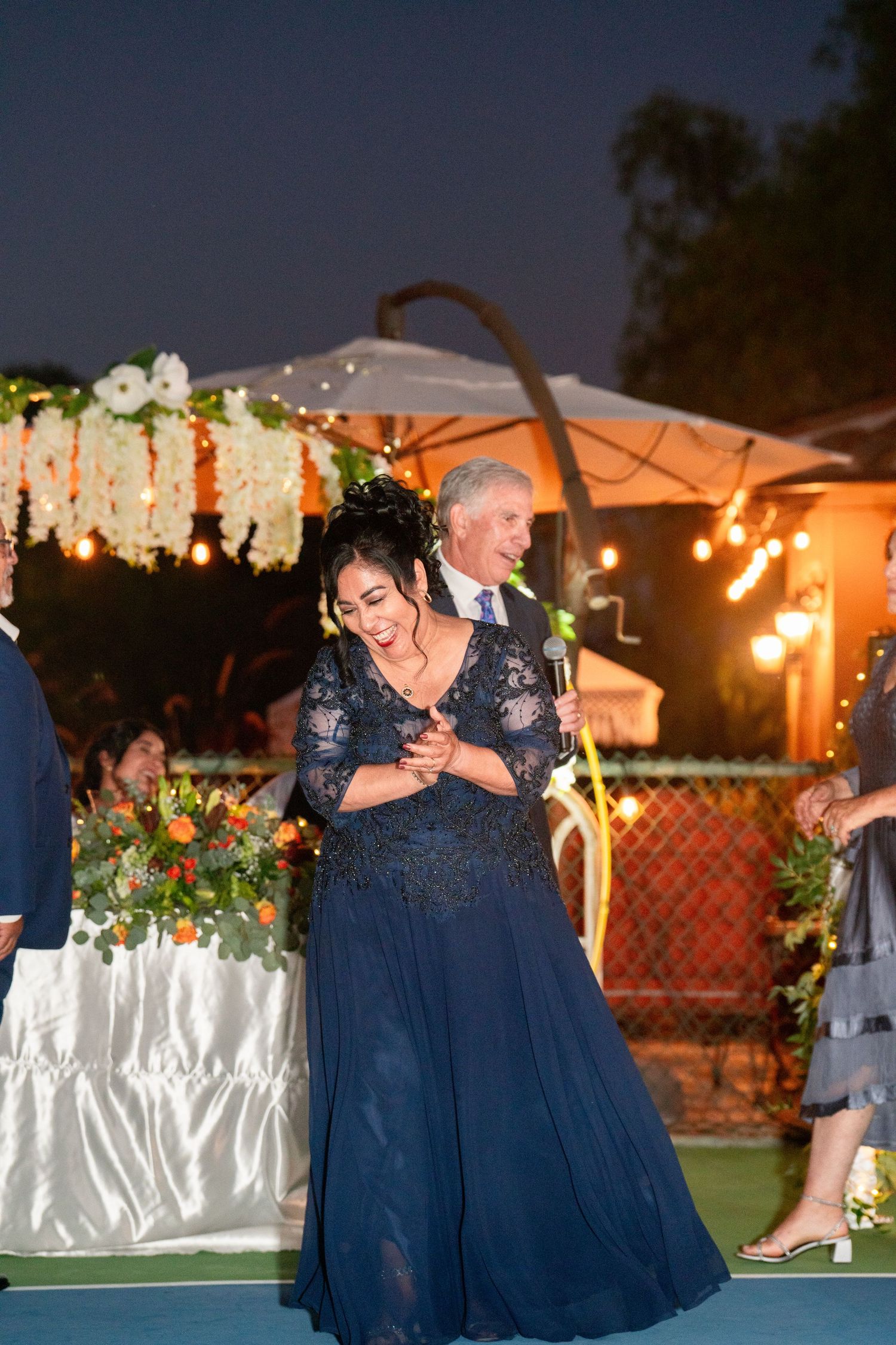 A woman in a navy evening gown dances under twinkling lights at an outdoor evening reception with floral decorations.