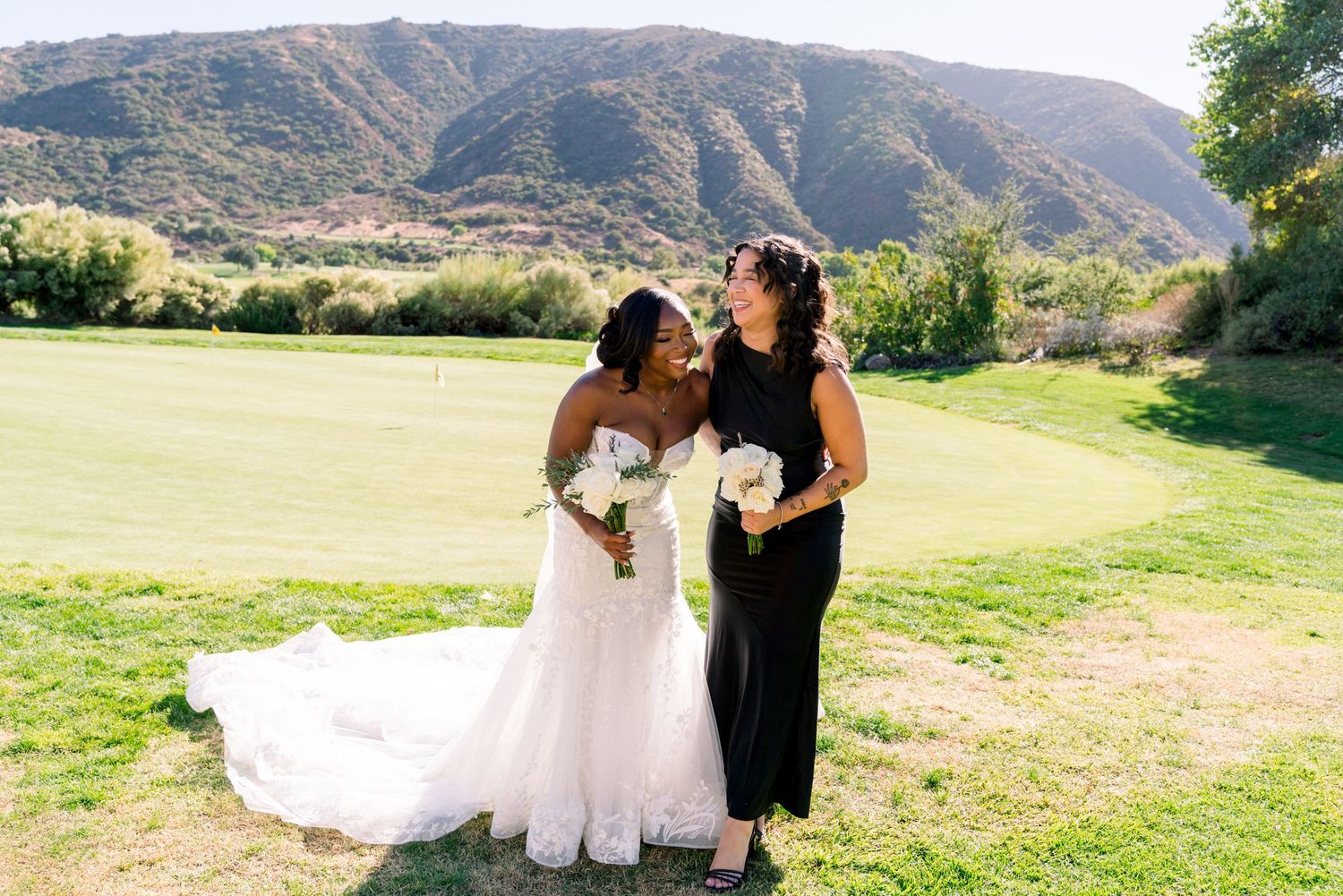 Two people in wedding attire sharing a moment on a grassy field with mountains in background.