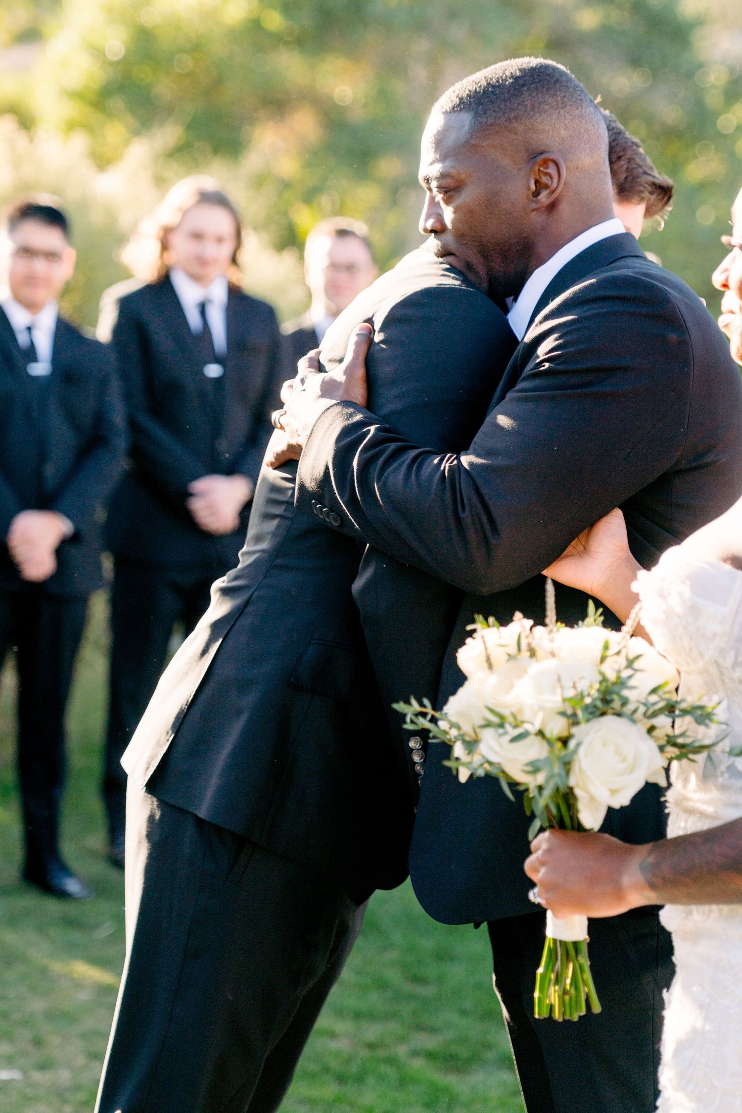 Emotional embrace between two men in black suits during outdoor wedding ceremony with bridal bouquet visible.