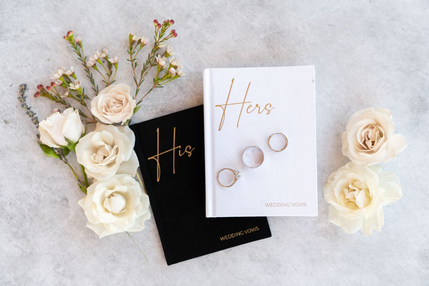 His and Hers wedding cards with gold lettering displayed with wedding rings and cream roses.