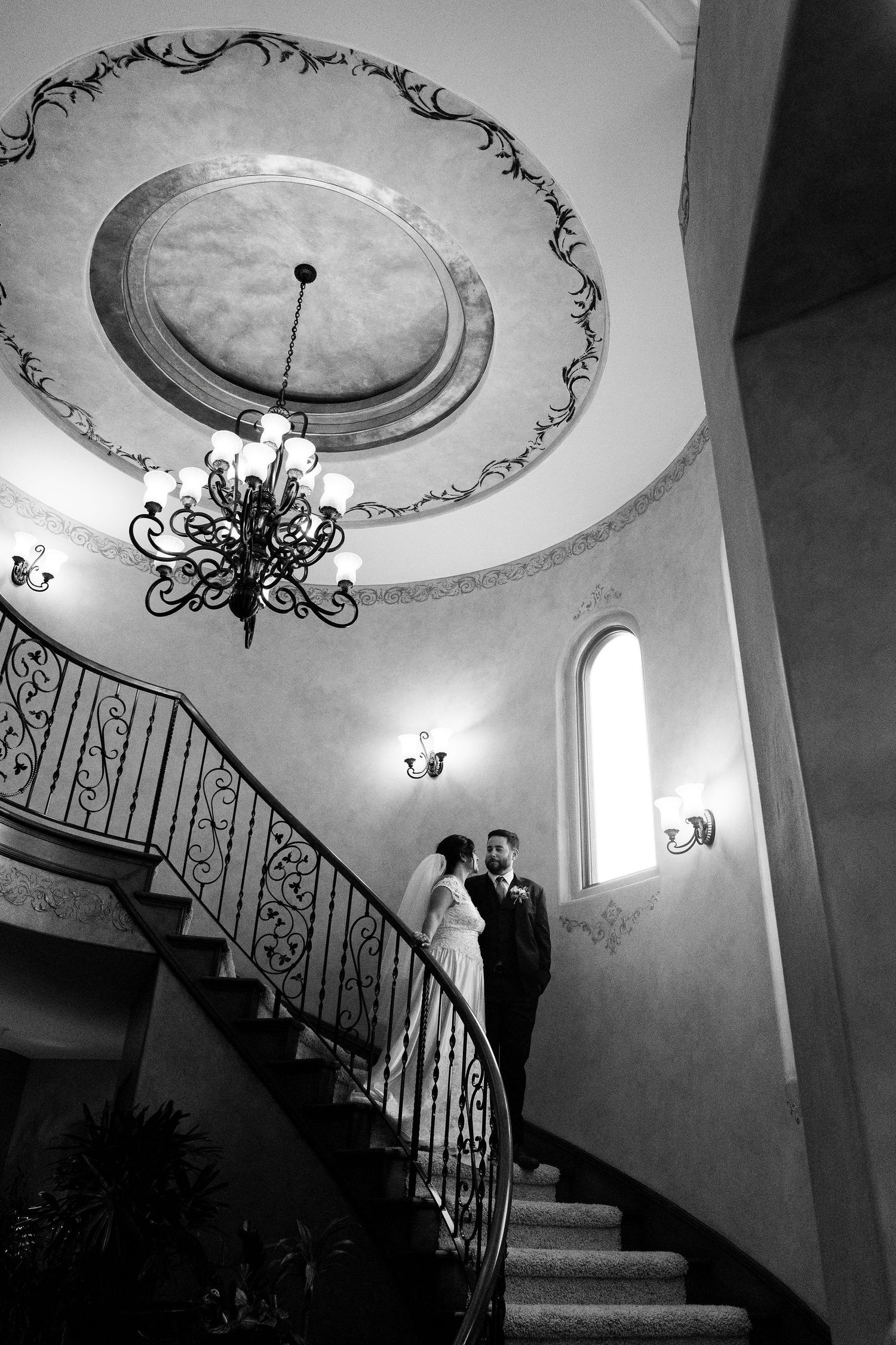 Elegant black and white architectural photo of a curved staircase with ornate ceiling medallion and chandelier in a formal setting.