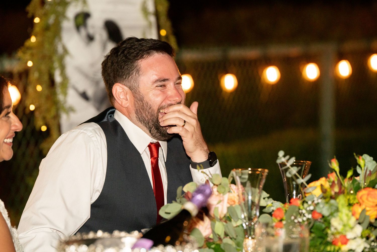 Wedding guest laughs while sitting at decorated reception table with string lights overhead.