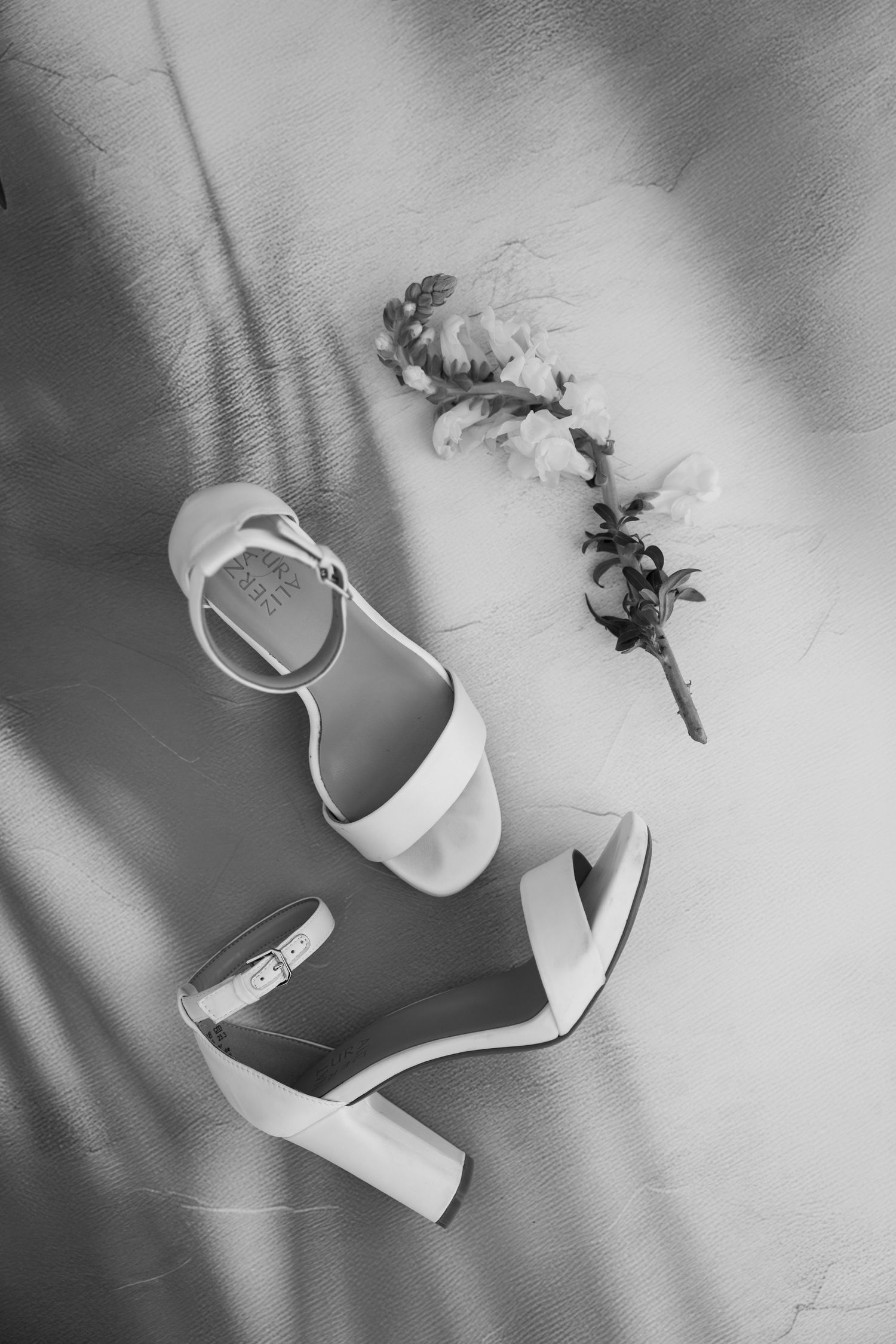 Elegant white wedding heels and dried flowers arranged on a light background with dramatic shadows.