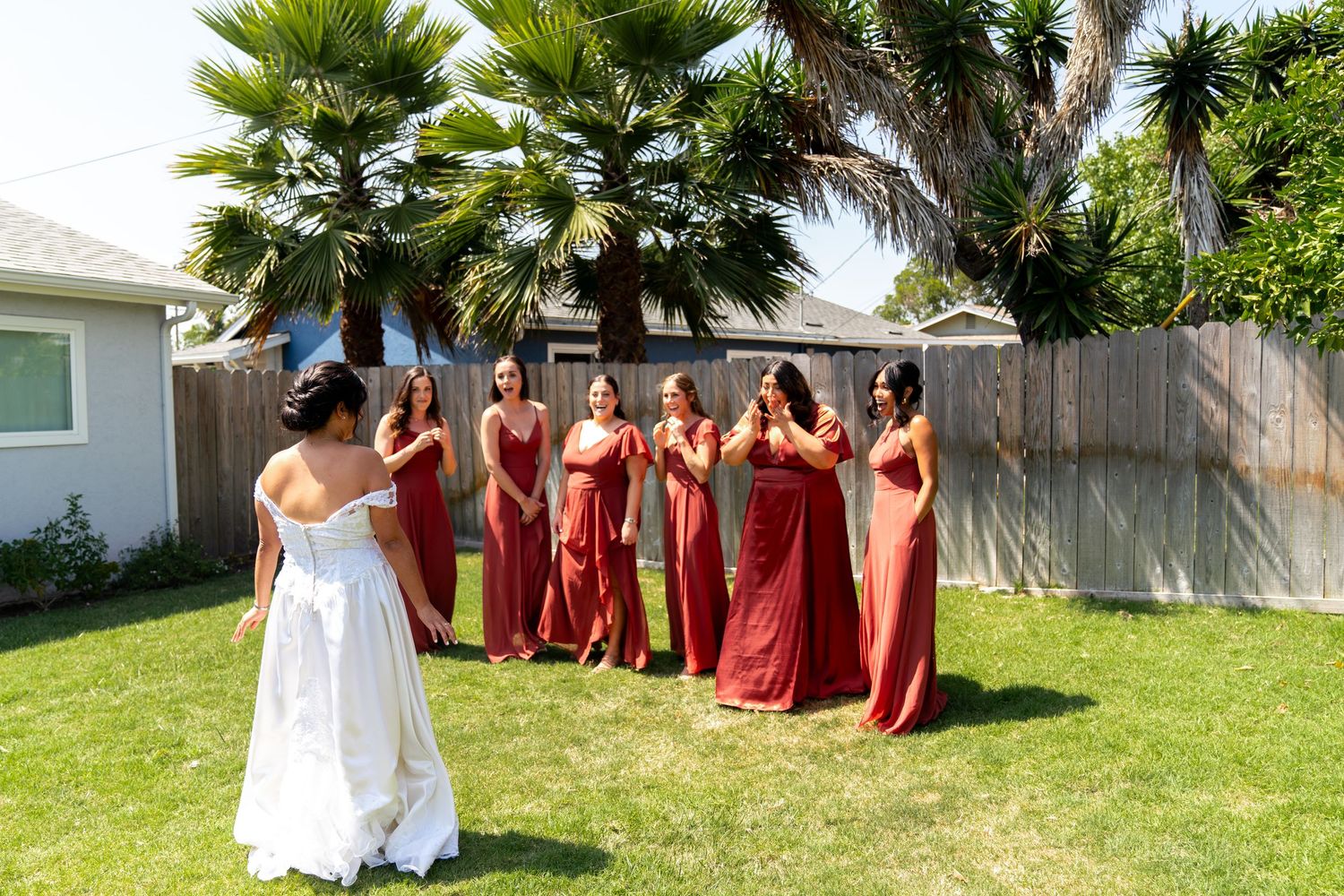 Wedding party wearing burgundy dresses reacts joyfully to the bride in a backyard setting with palm trees.