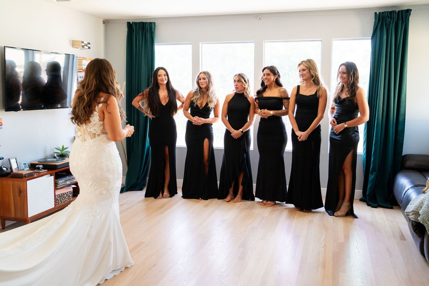 Bridesmaids in matching black dresses react emotionally as they see the bride in her white gown in a sunlit room.