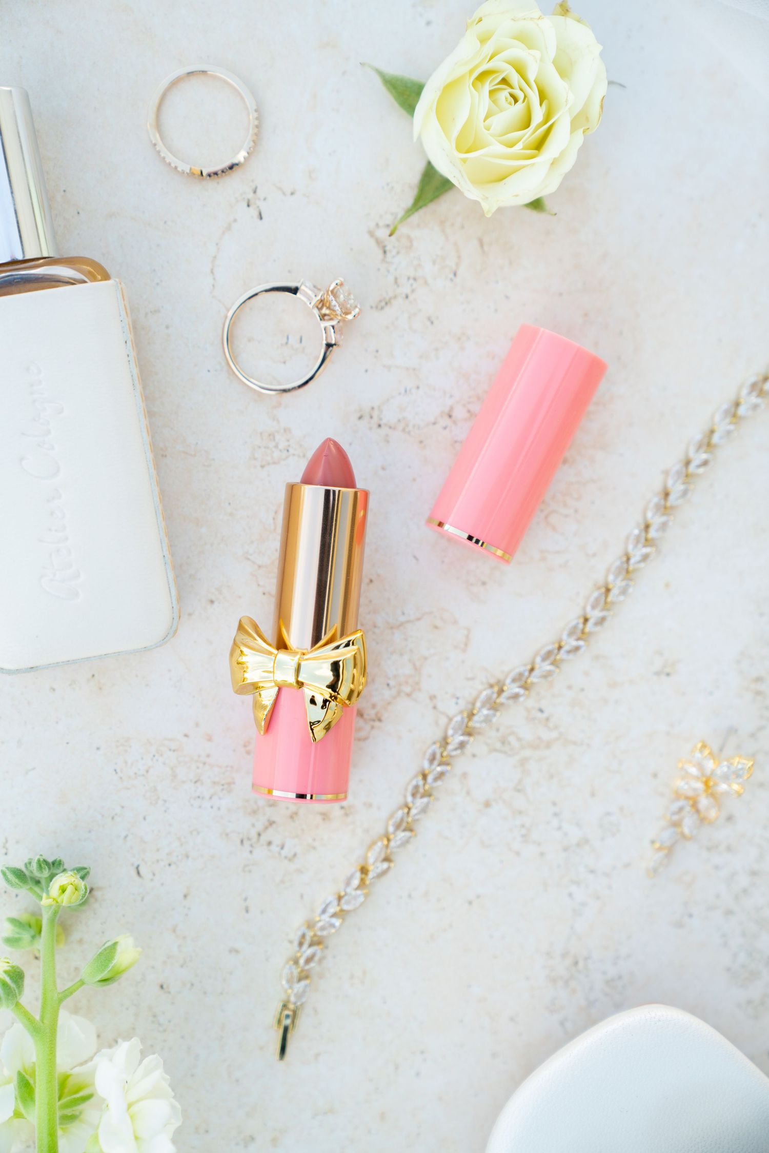 Pink lipstick with gold bow detail alongside rings, white roses and feminine accessories on light marble surface.