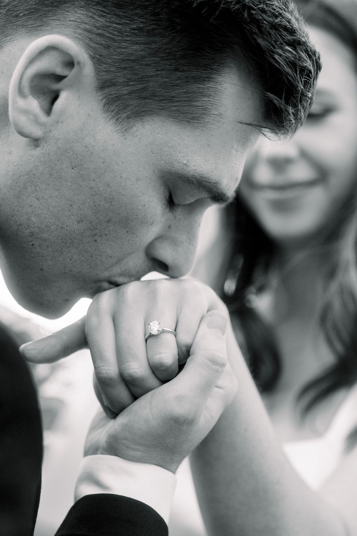 Black and white romantic image showing a groom kissing a wedding ring with soft focus on a blurred bride in background.