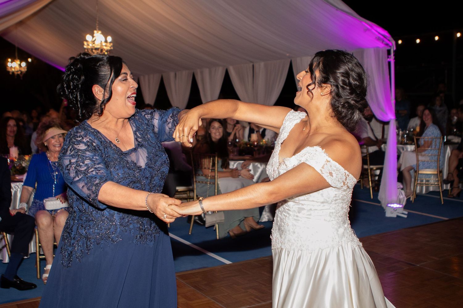 A joyful dance moment between a bride and another guest under an elegant tented reception with purple lighting.